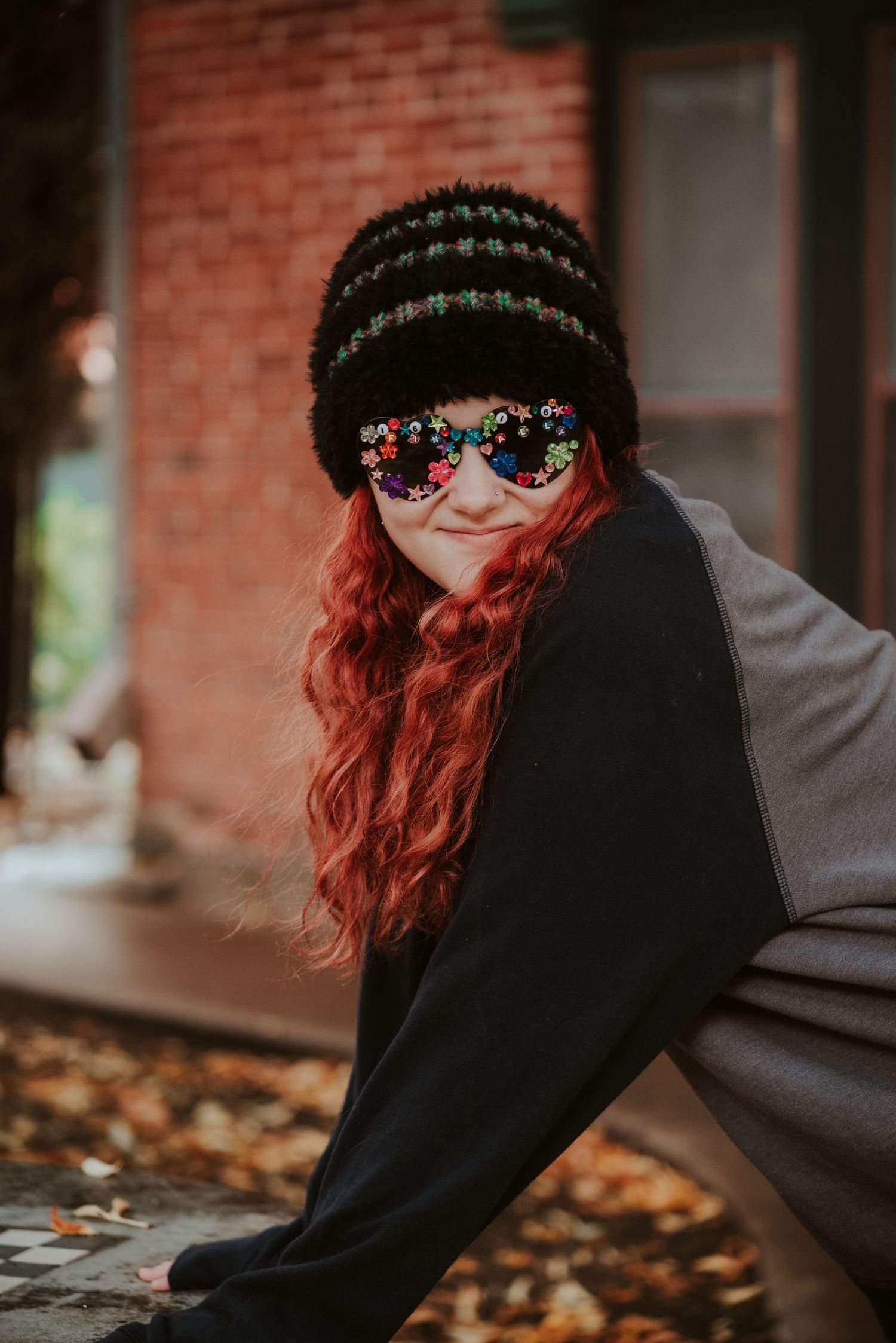 Woman with long red hair wearing a black beanie and floral sunglasses posing outdoors in autumn.