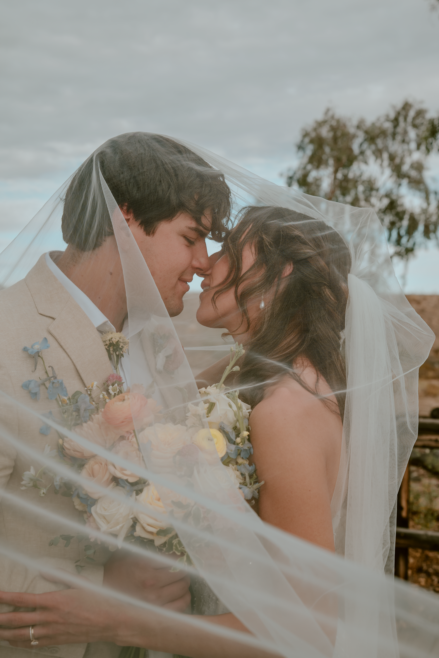 A bride and groom share an intimate moment under a flowing wedding veil outdoors on their wedding day.