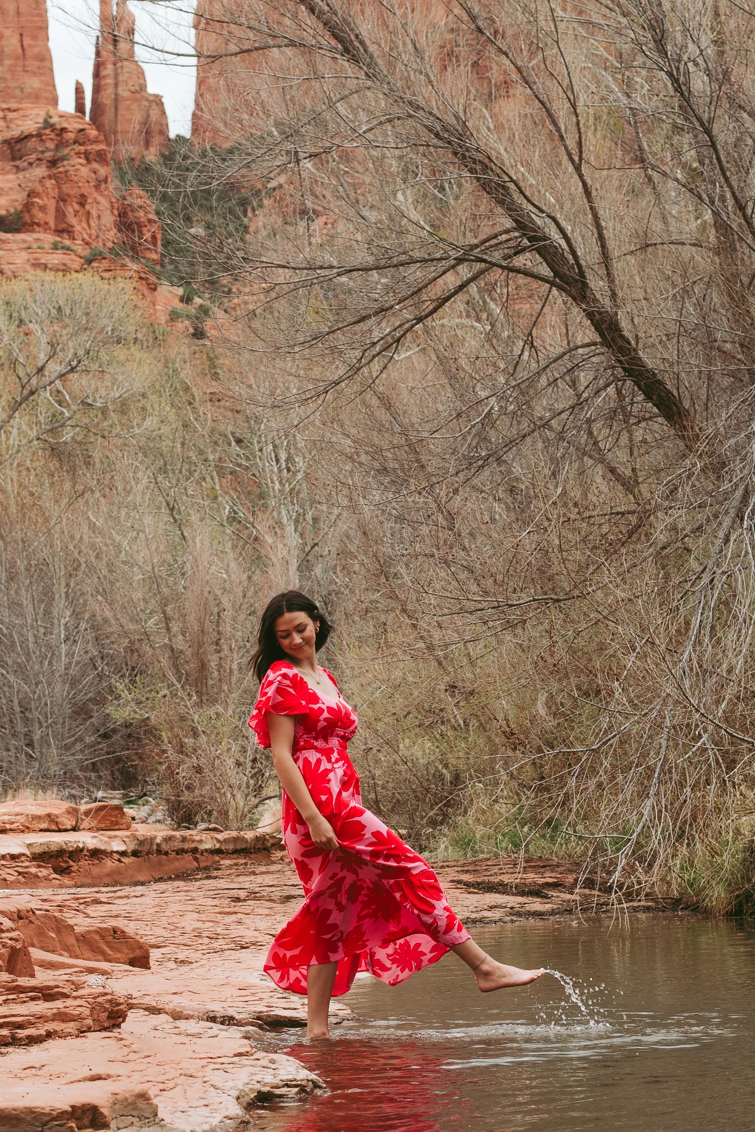 Red dress flowing gracefully while stepping in water near red rock cliffs.