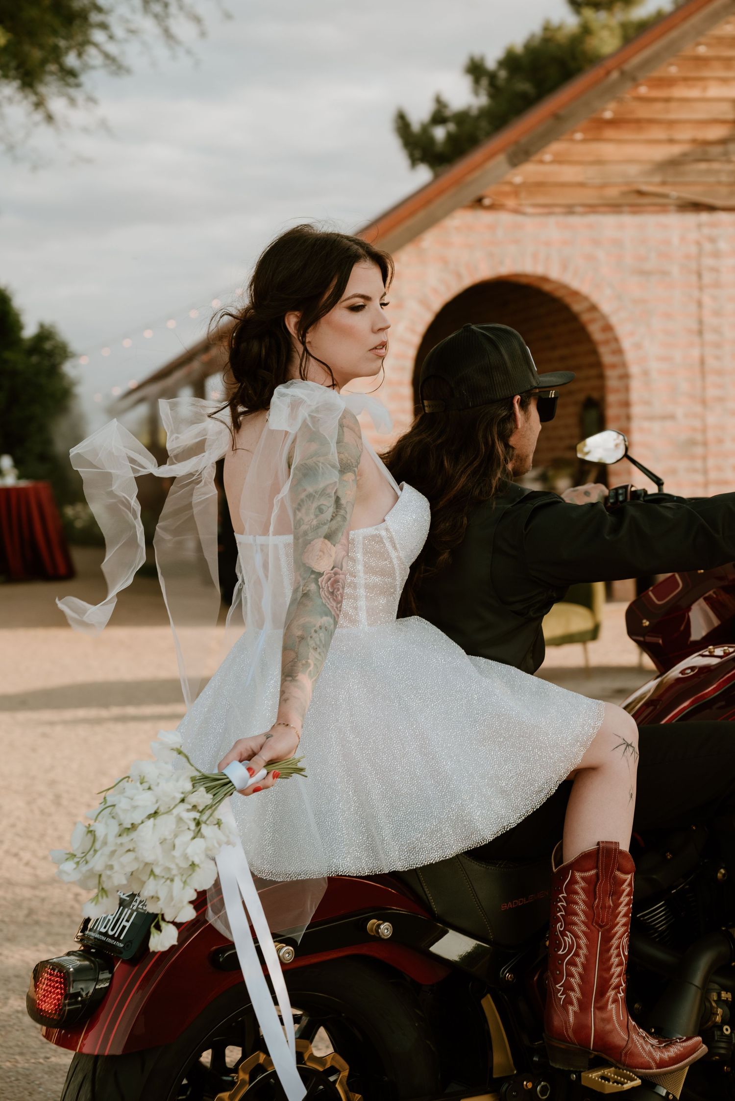 Bride in white dress and cowboy boots posed with vintage motorcycle.