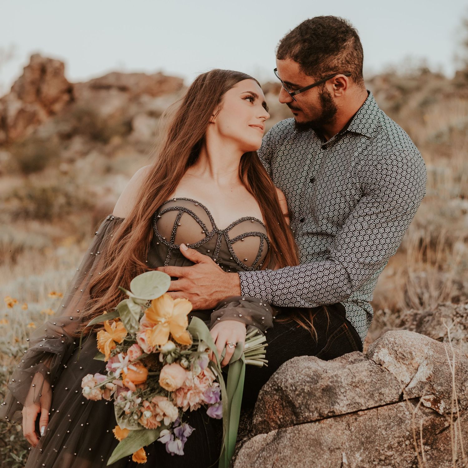 A couple poses intimately on desert rocks with a bouquet of pastel flowers.