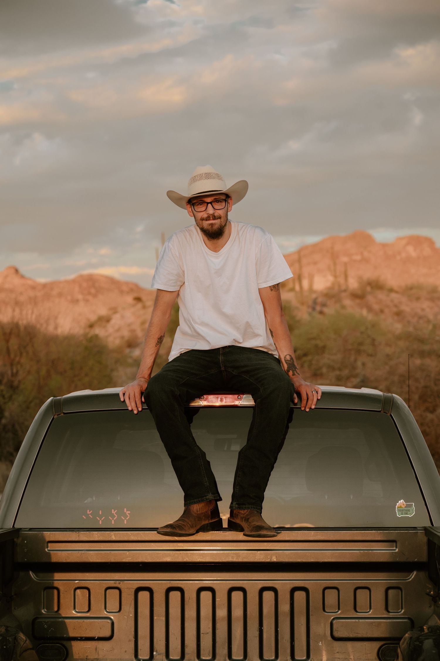 Man in cowboy hat and white tee sits on truck hood in desert landscape at golden hour.