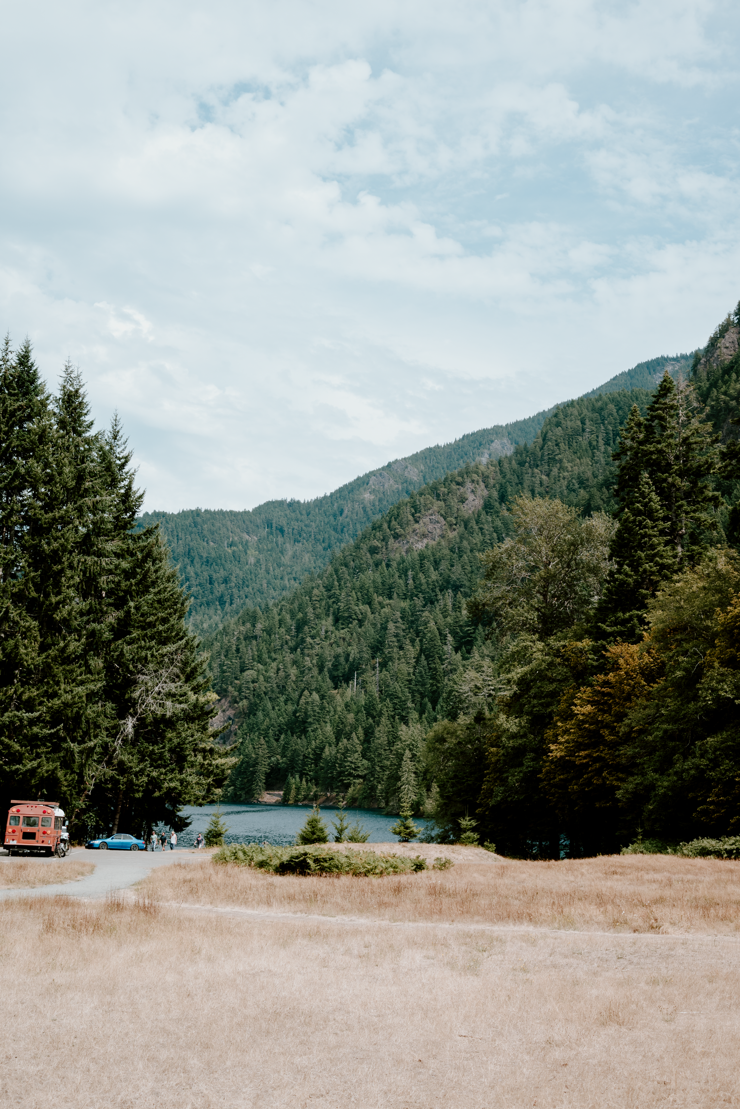A serene mountain landscape with a small lake, dense evergreen forest, dry meadow, and a red vehicle under a partly cloudy sky.