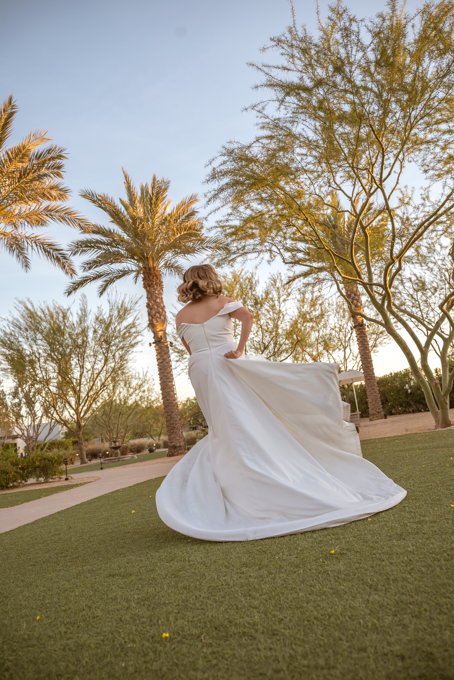 Bride in white off-shoulder gown twirling on green lawn with palm trees at golden hour.
