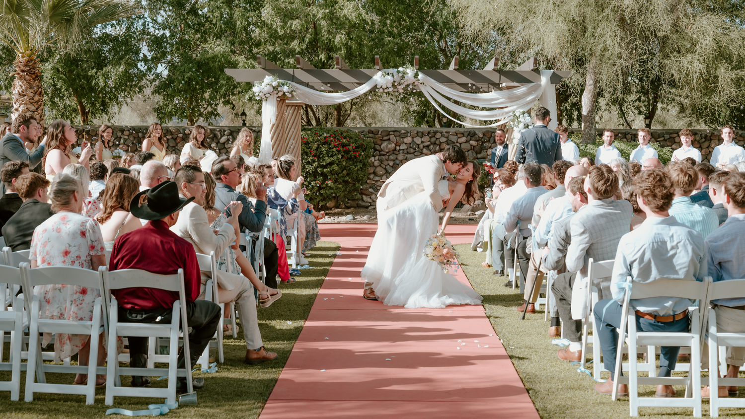 Outdoor wedding ceremony with bride and groom kissing at altar on red aisle, guests seated in white chairs under trees.