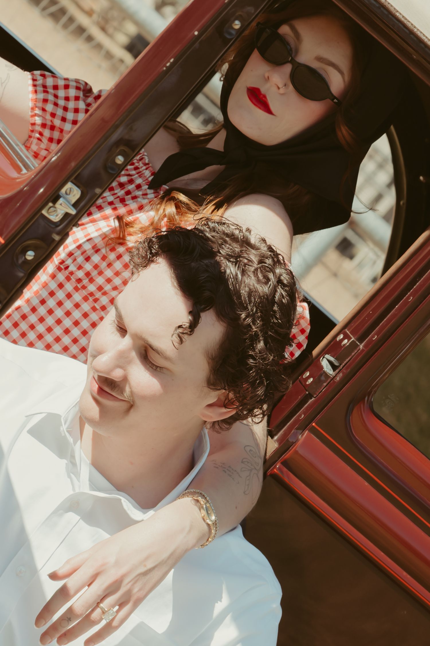 A retro-styled couple near a red vintage car, woman in sunglasses with red lips leaning over a curly-haired man.