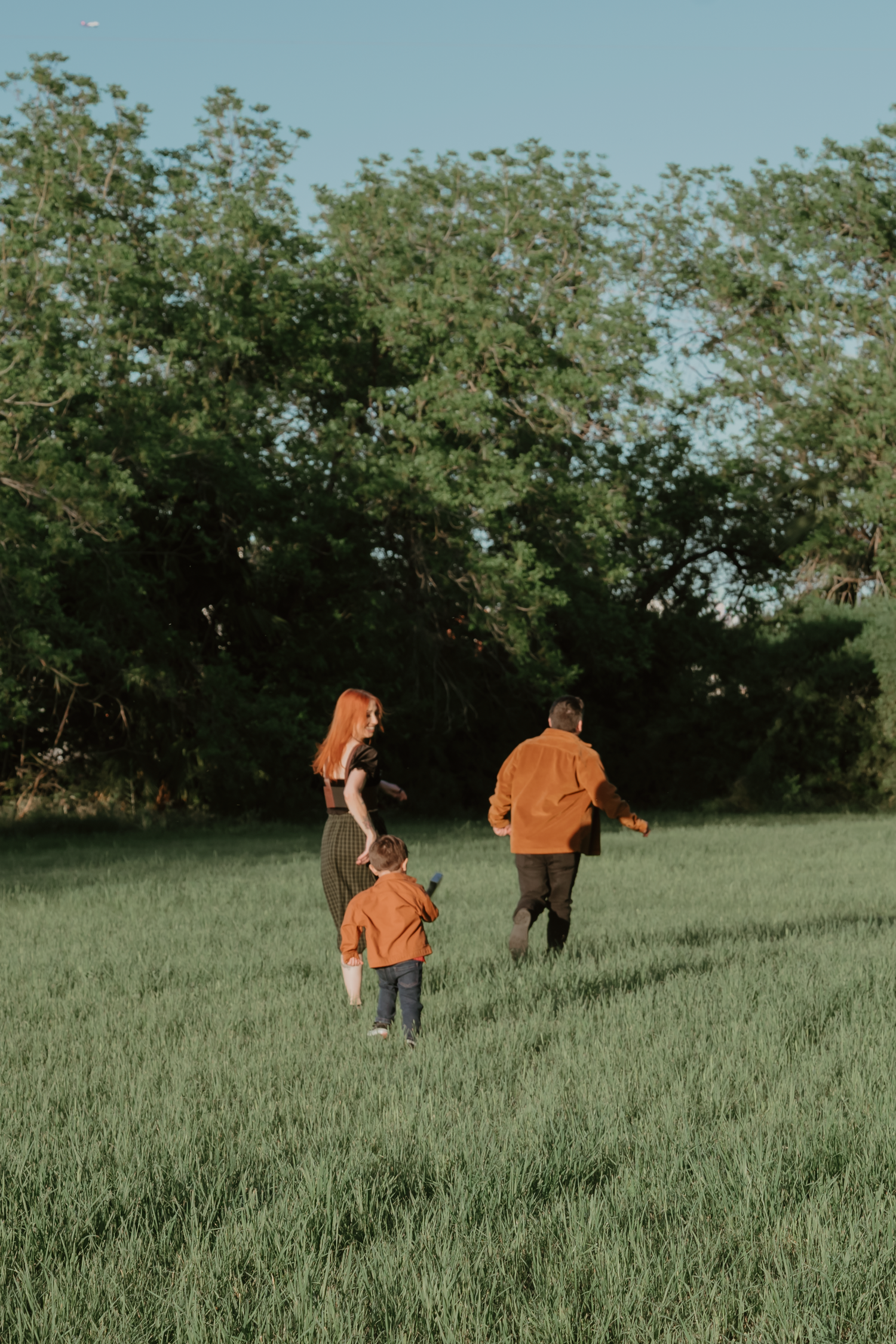 Family of three walking through green field toward trees at golden hour, viewed from behind.