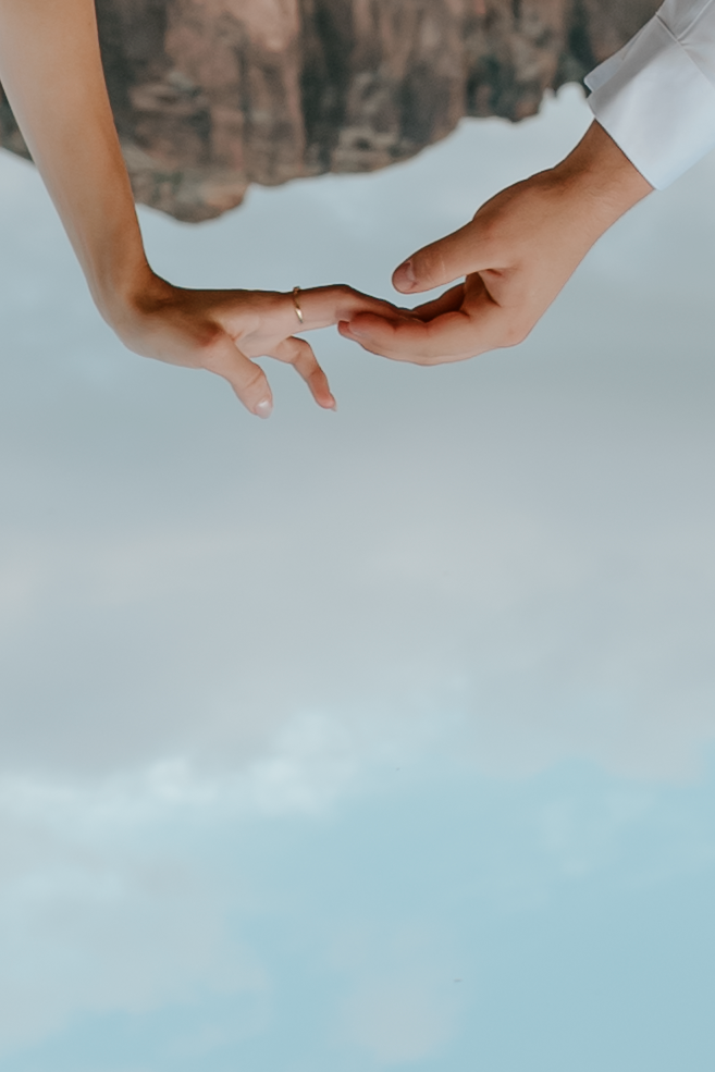 Two hands gently touching fingertips with an engagement ring visible, set against a soft blue sky backdrop.