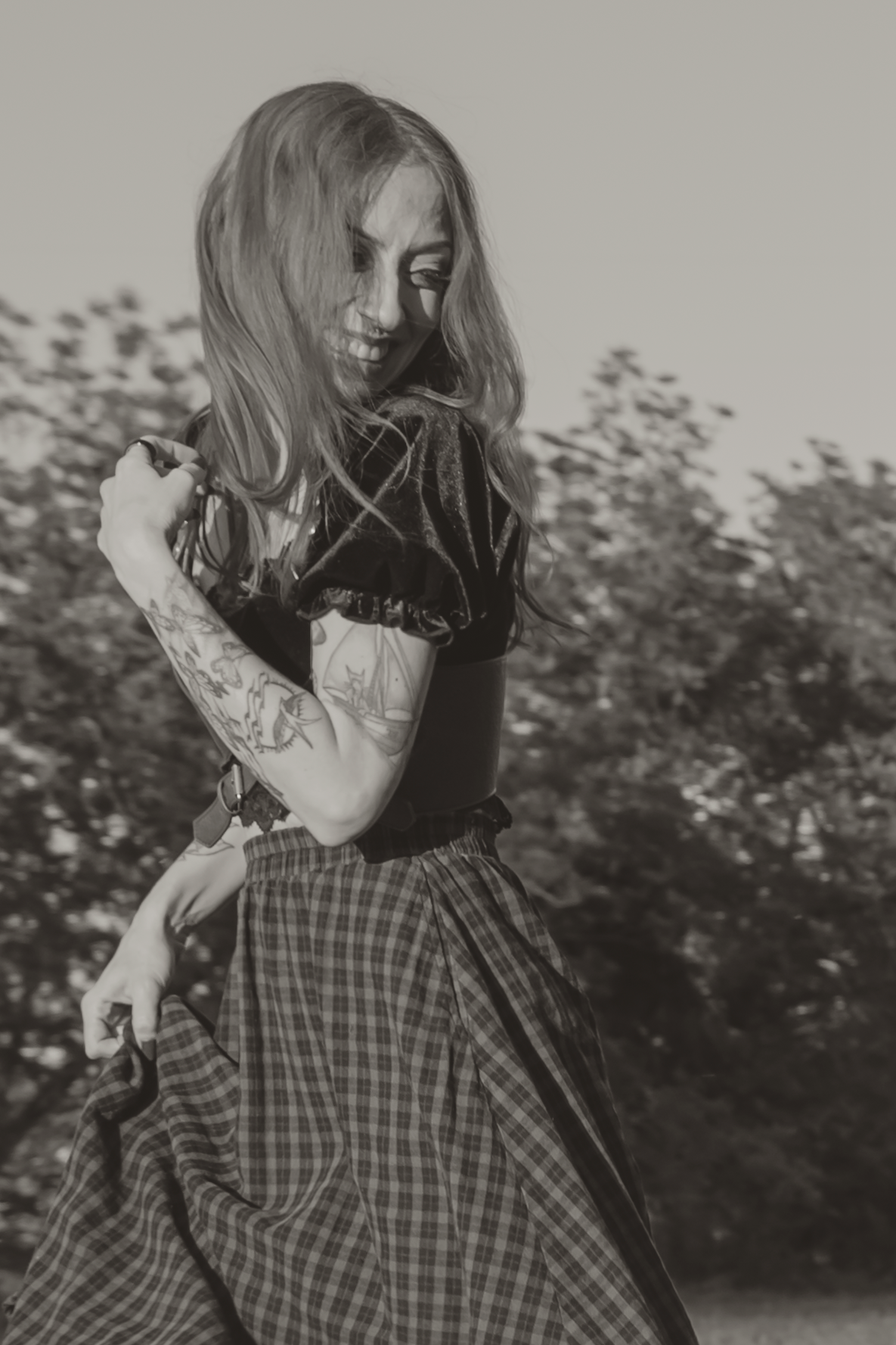 Black and white photo of a tattooed woman with long hair wearing a plaid skirt, posing outdoors near trees.