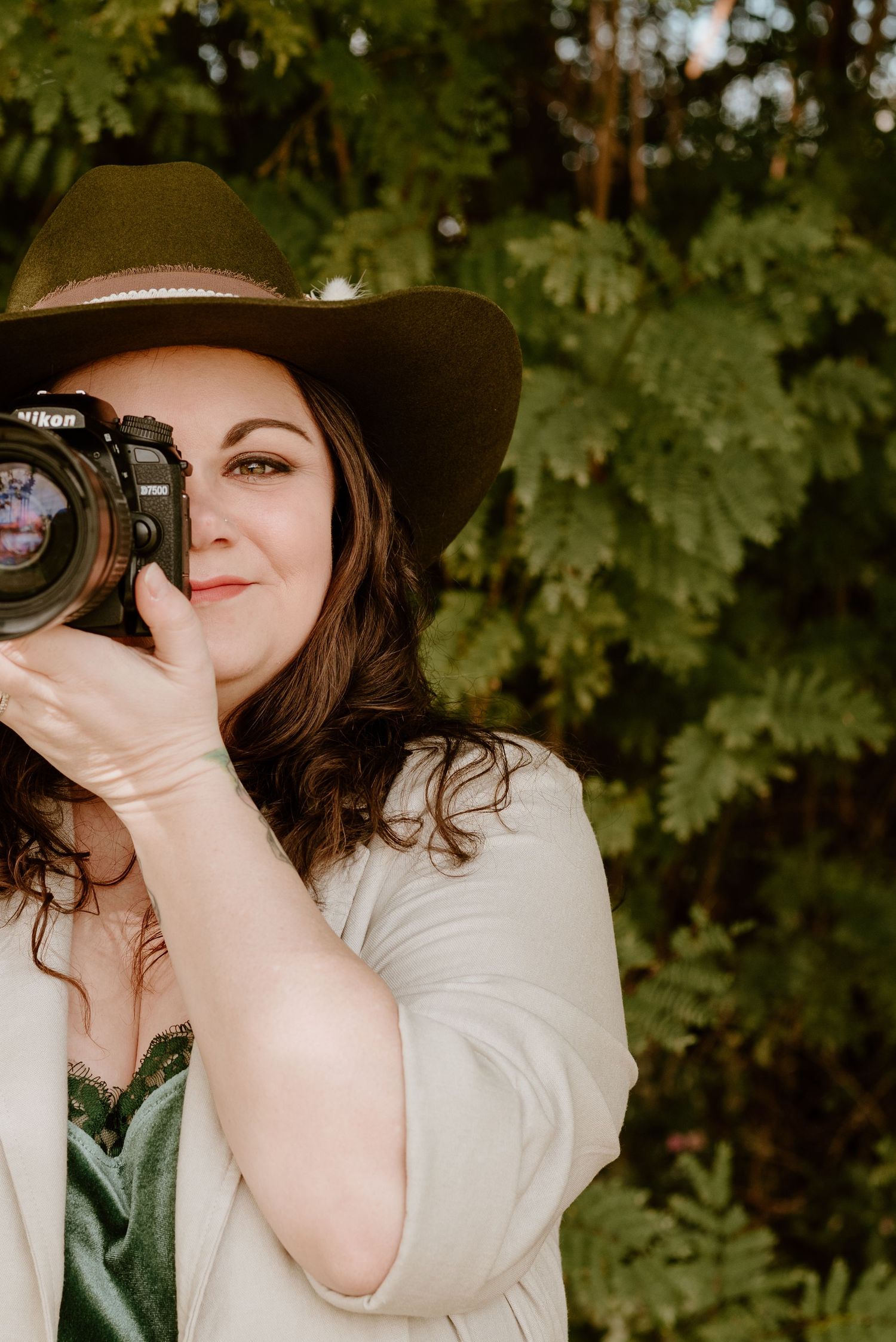 A photographer in a brown hat uses a professional camera against a leafy green background.