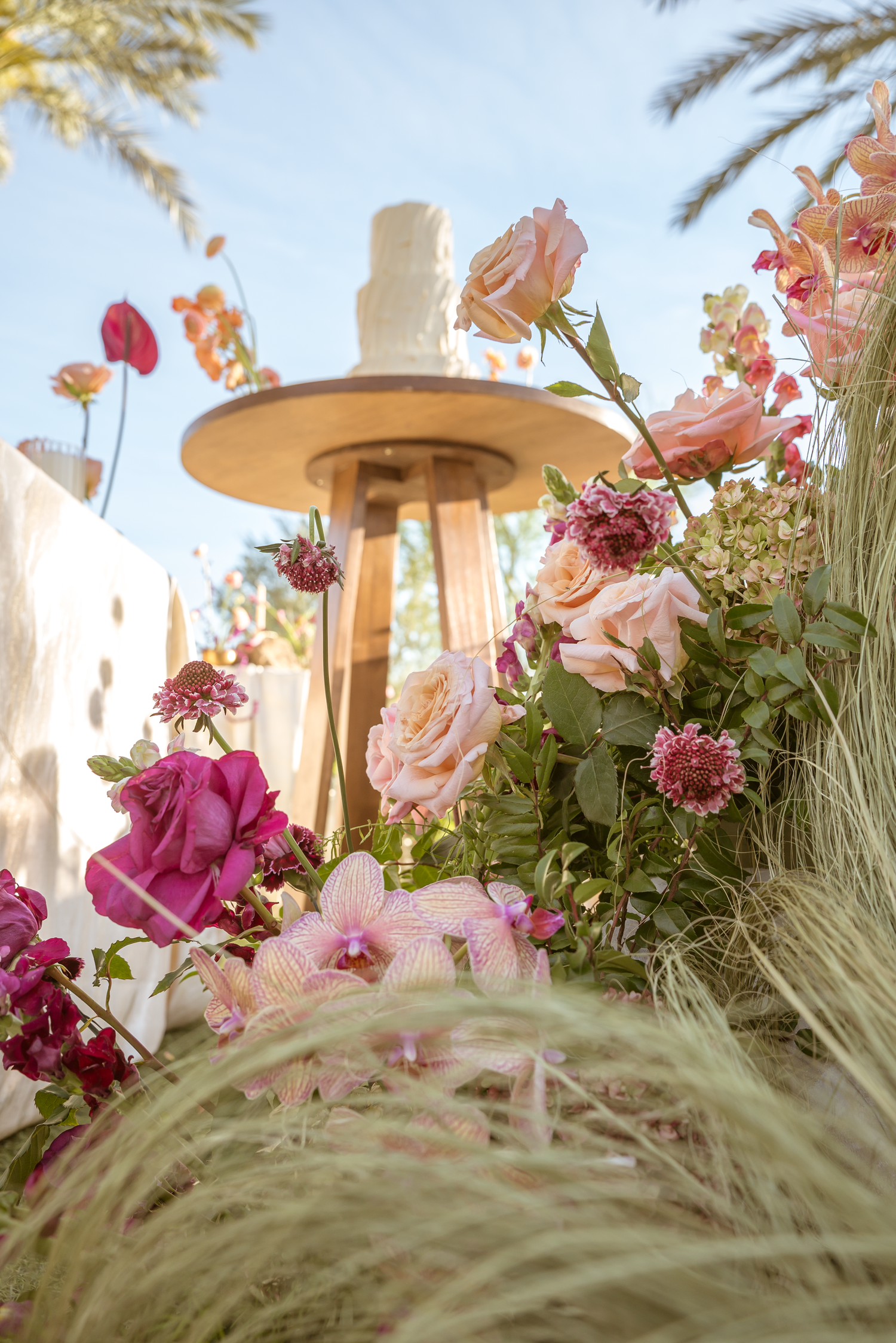 White wedding cake on wooden stand surrounded by lush pink roses, orchids, and wildflowers outdoors.