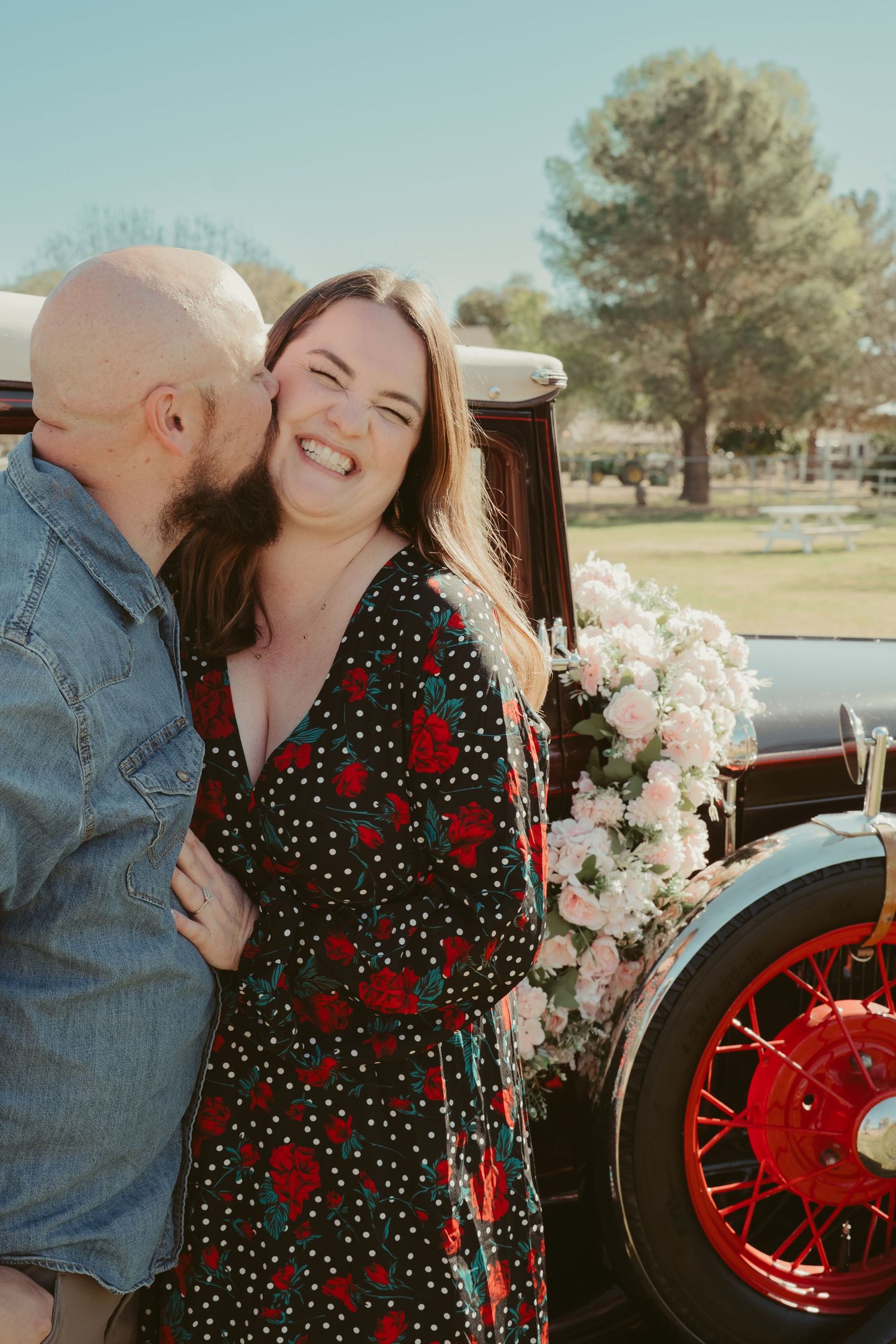 A couple shares a romantic moment by a vintage car adorned with flowers, as the man kisses the laughing woman's cheek.