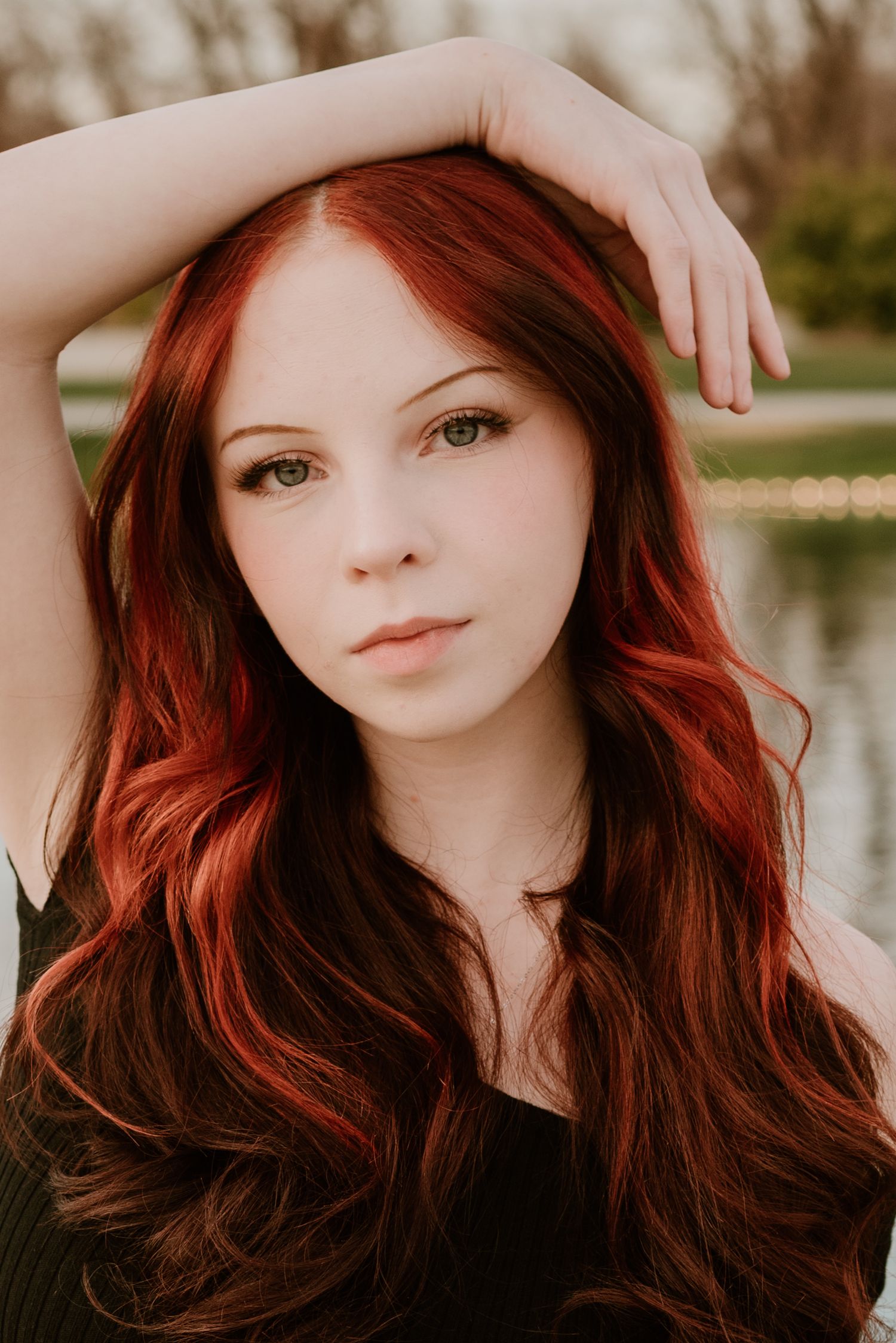 Portrait with flowing red hair and dramatic makeup against a lakeside backdrop.