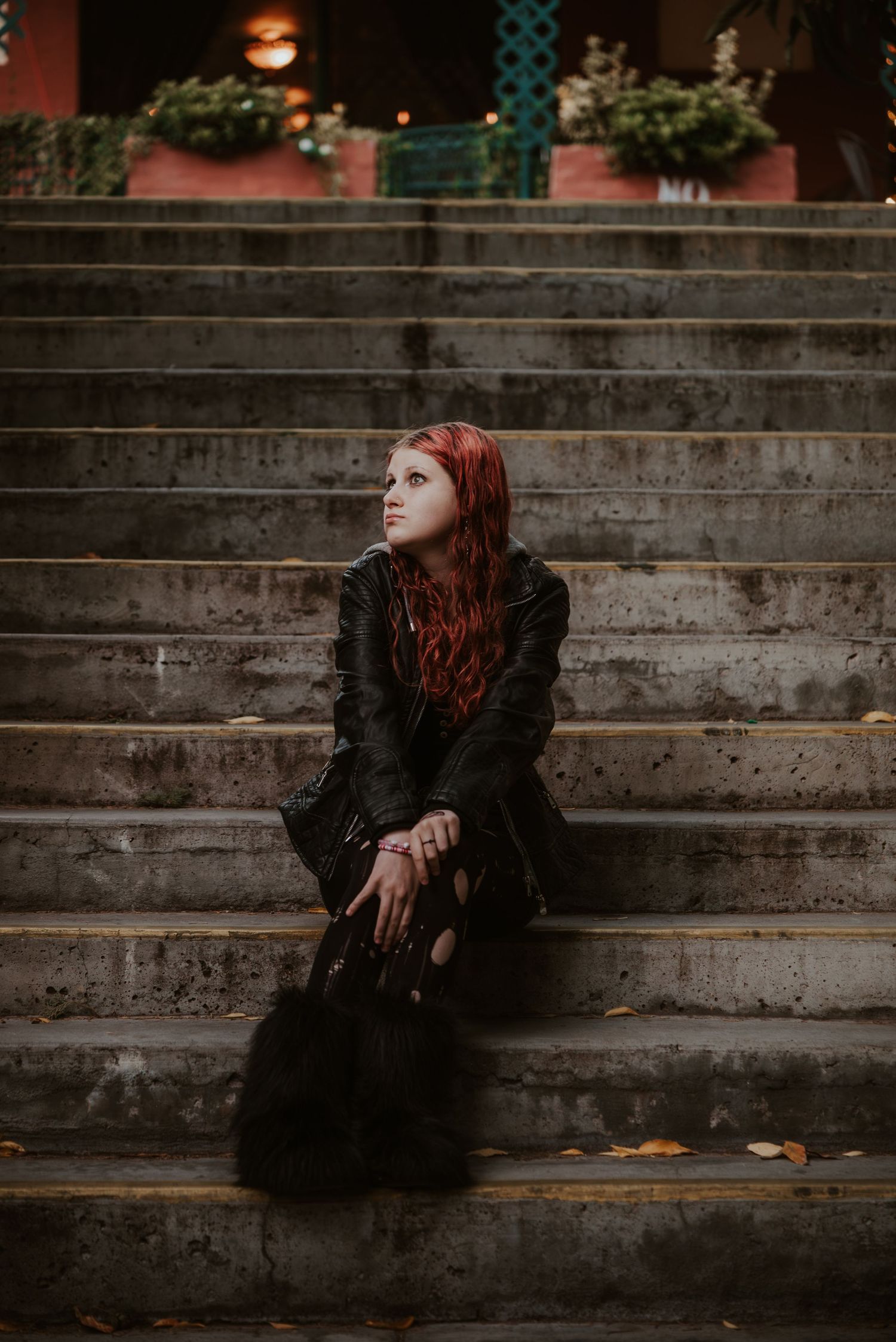A woman with red hair sits on concrete steps wearing a black leather jacket and dark flowy skirt, gazing upward.