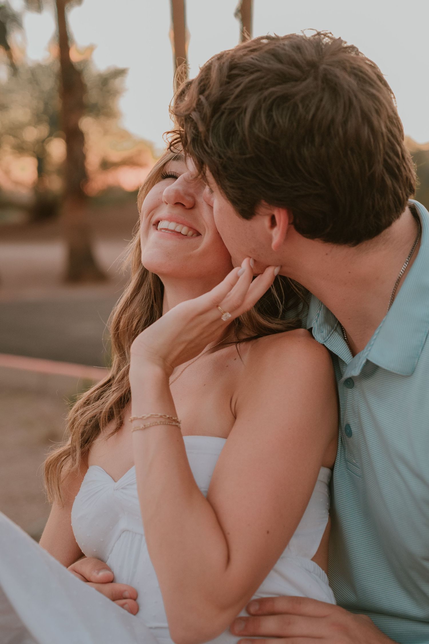 A smiling bride in a white dress receives a cheek kiss from her groom in a light blue shirt during a golden hour engagement session.