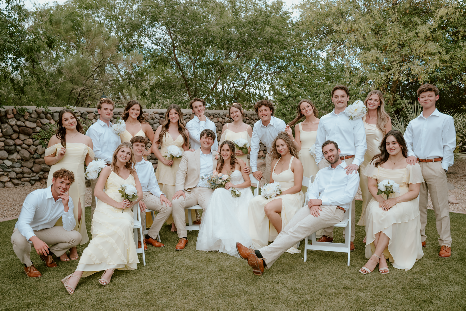 Large wedding party posing outdoors in neutral tones, bride and groom centered among smiling guests on green lawn.