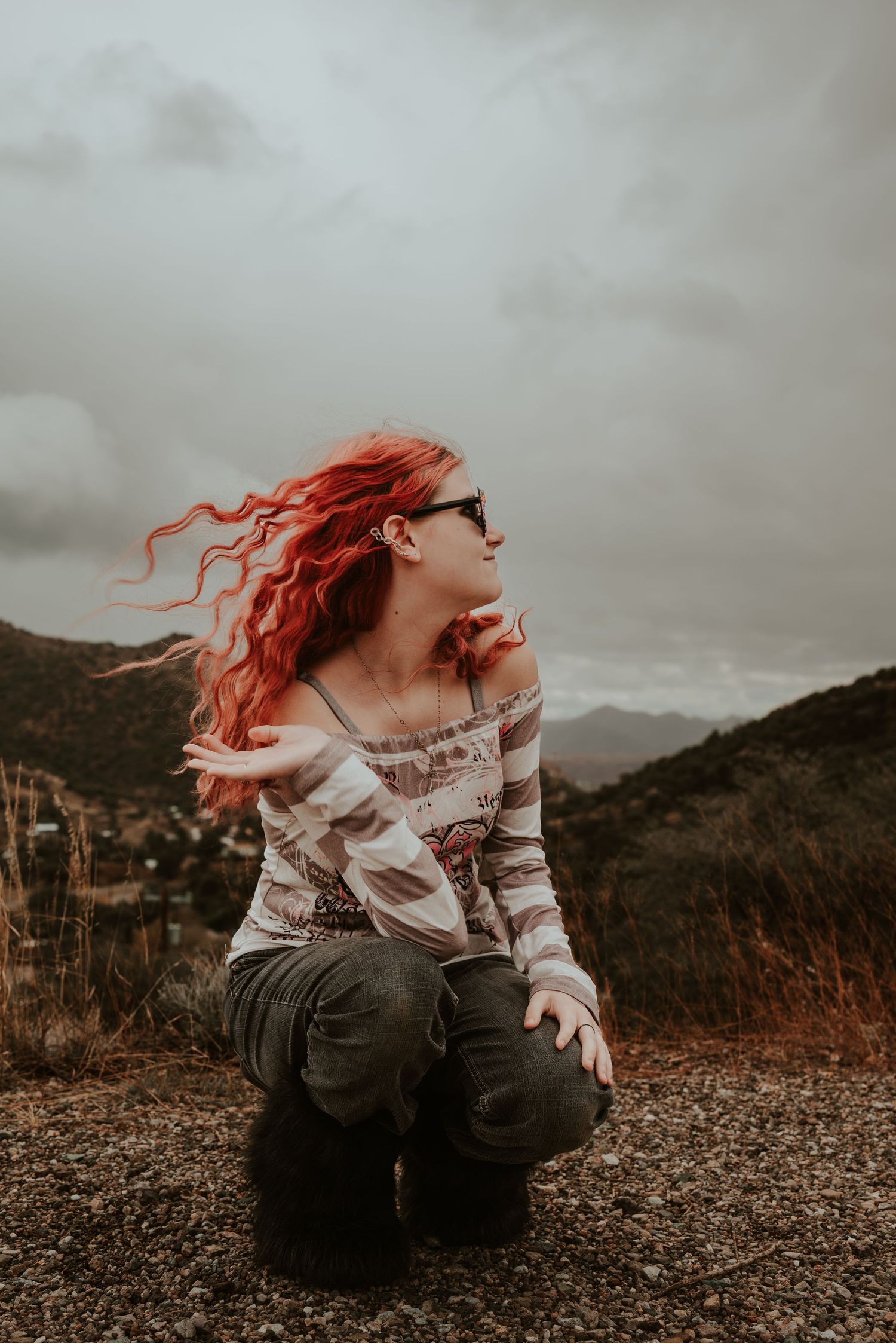 A woman with vibrant red hair kneels on a hilltop, her hair blowing in the wind against a cloudy sky.