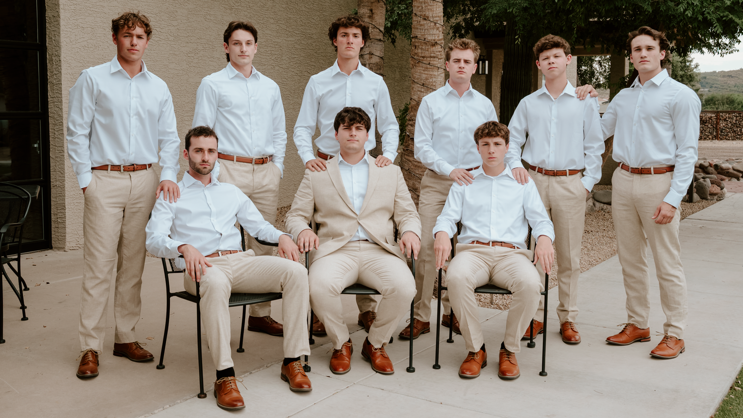 Nine young men in matching khaki pants and light blue shirts pose formally outdoors for a group wedding party photo.