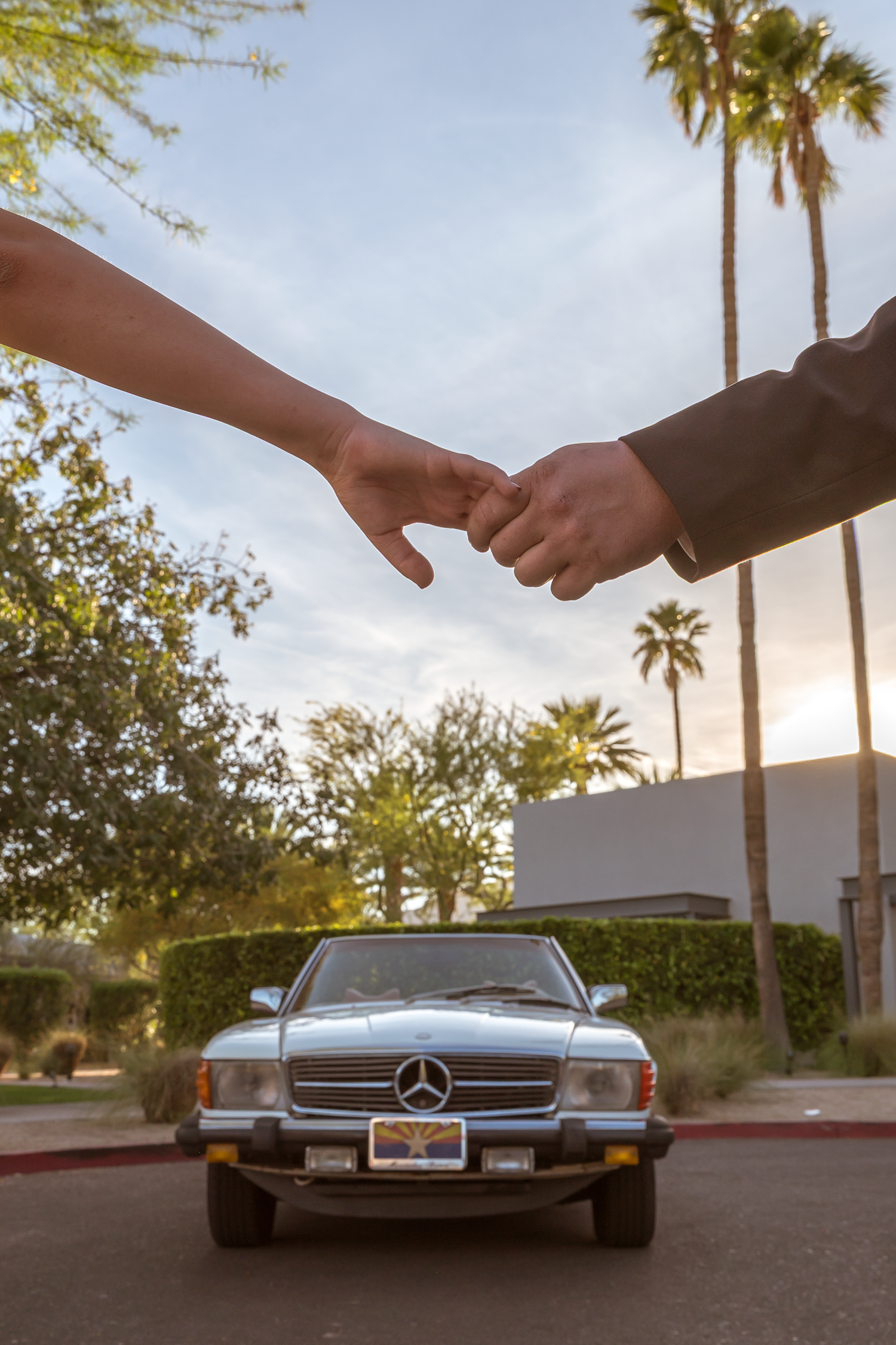 Two hands almost touching above a classic Mercedes-Benz convertible at sunset with palm trees in the background.