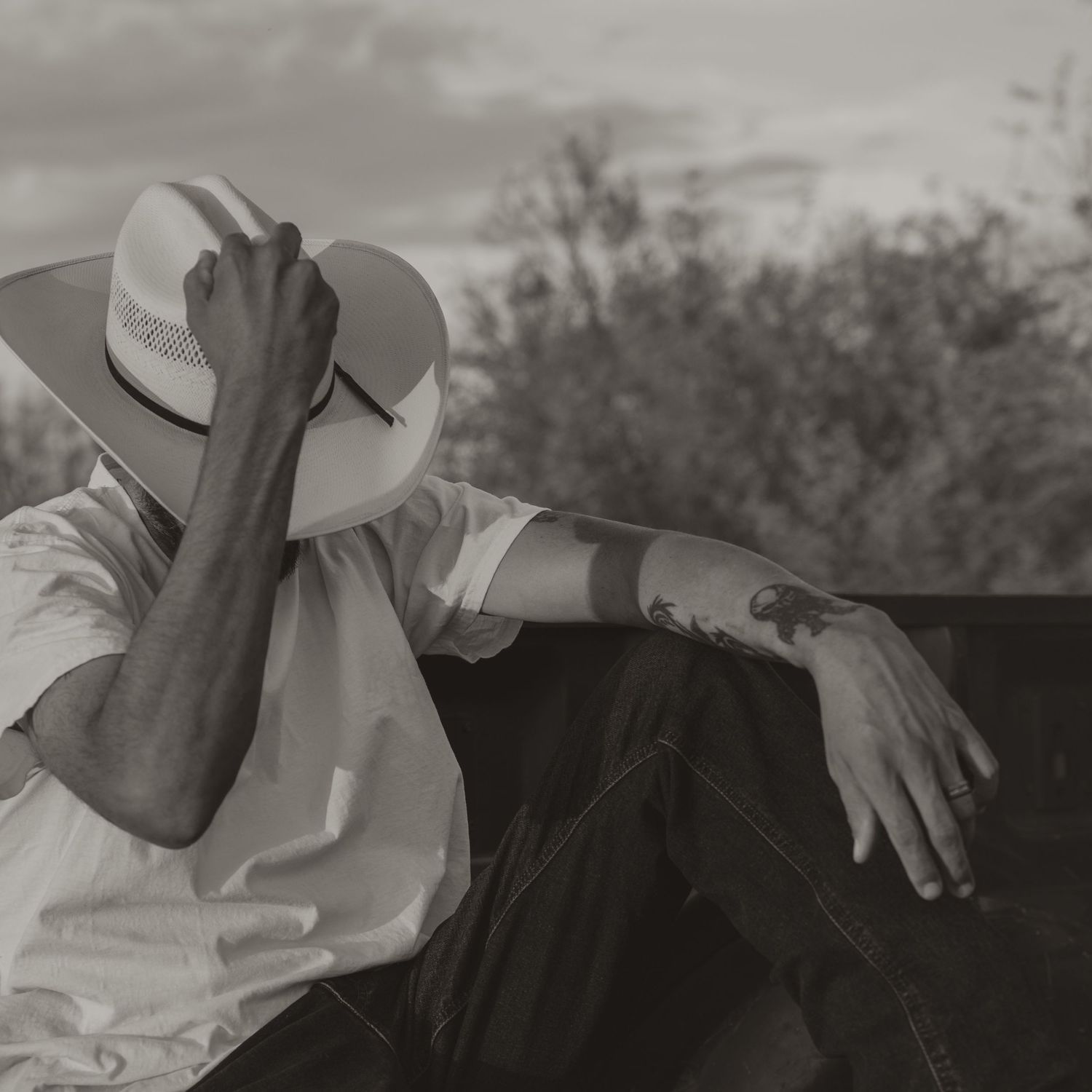 Black and white photo of a man in a white cowboy hat and tattoos sitting outdoors near trees.
