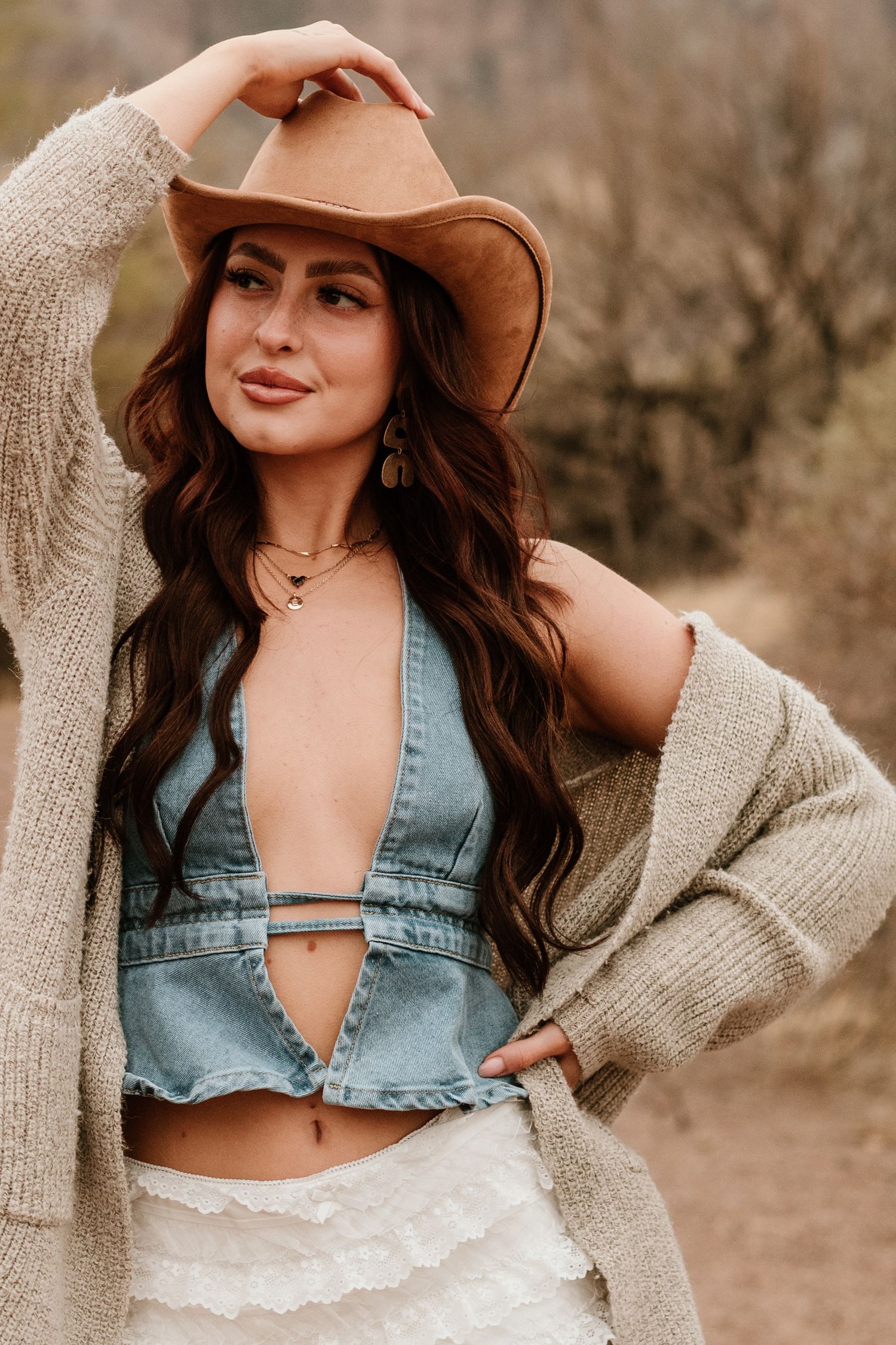 A stylish western-inspired look with denim, cardigan and brown hat against a natural backdrop.