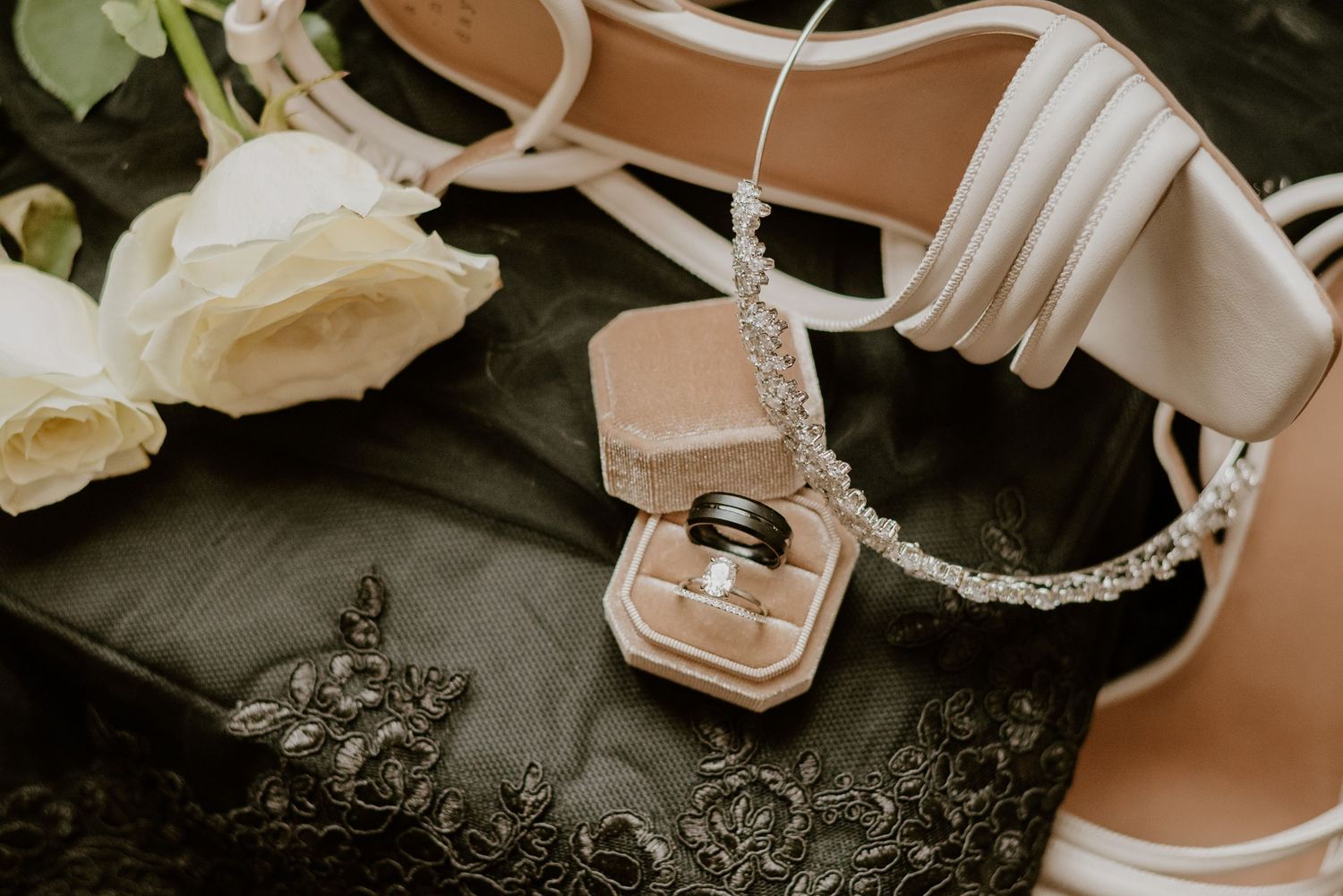 Wedding accessories including white sandals, roses, and a ring box arranged on dark fabric.
