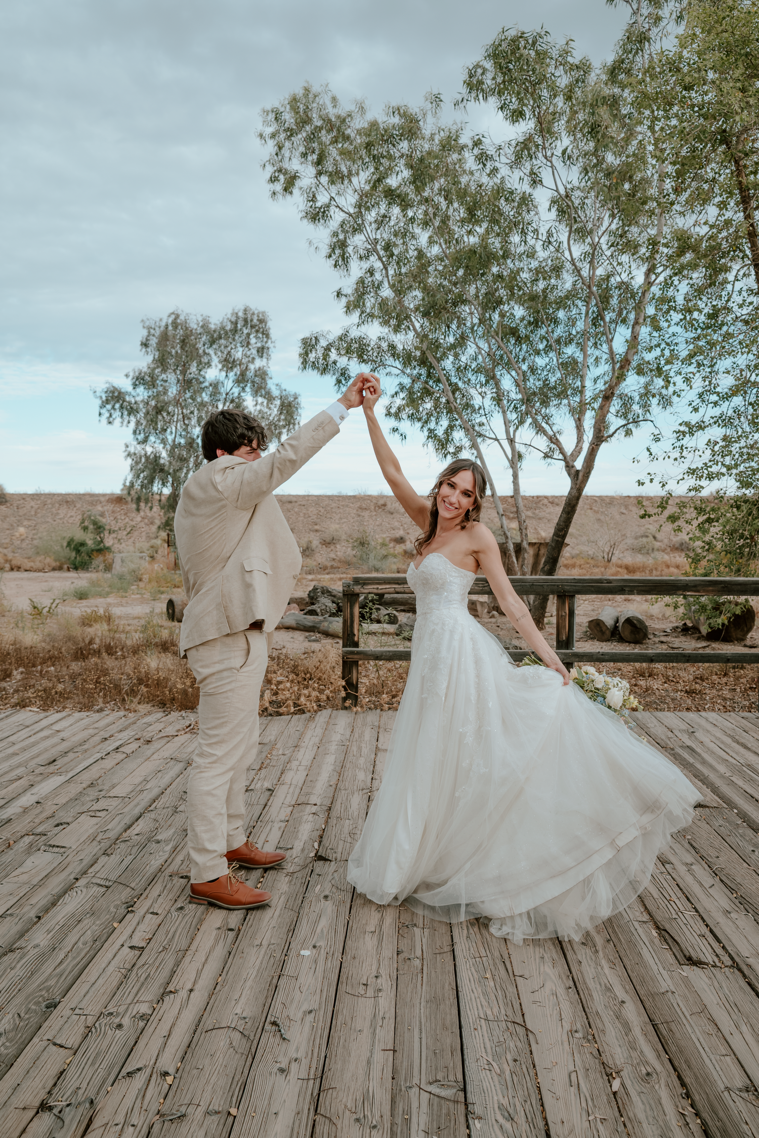 A bride in a white gown twirls with her groom in a tan suit on a wooden deck outdoors.