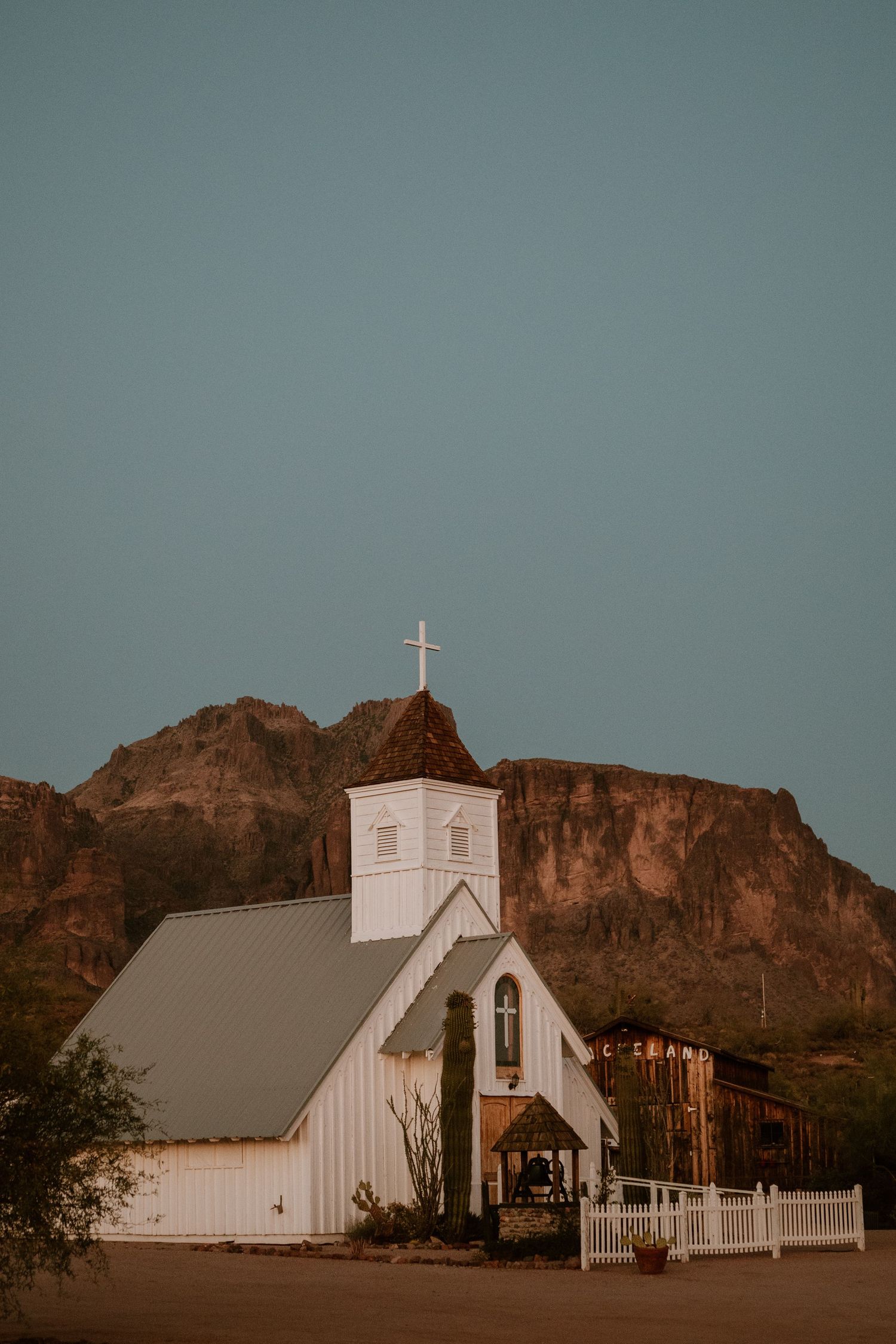 White church with cross against desert mountain backdrop at golden hour.