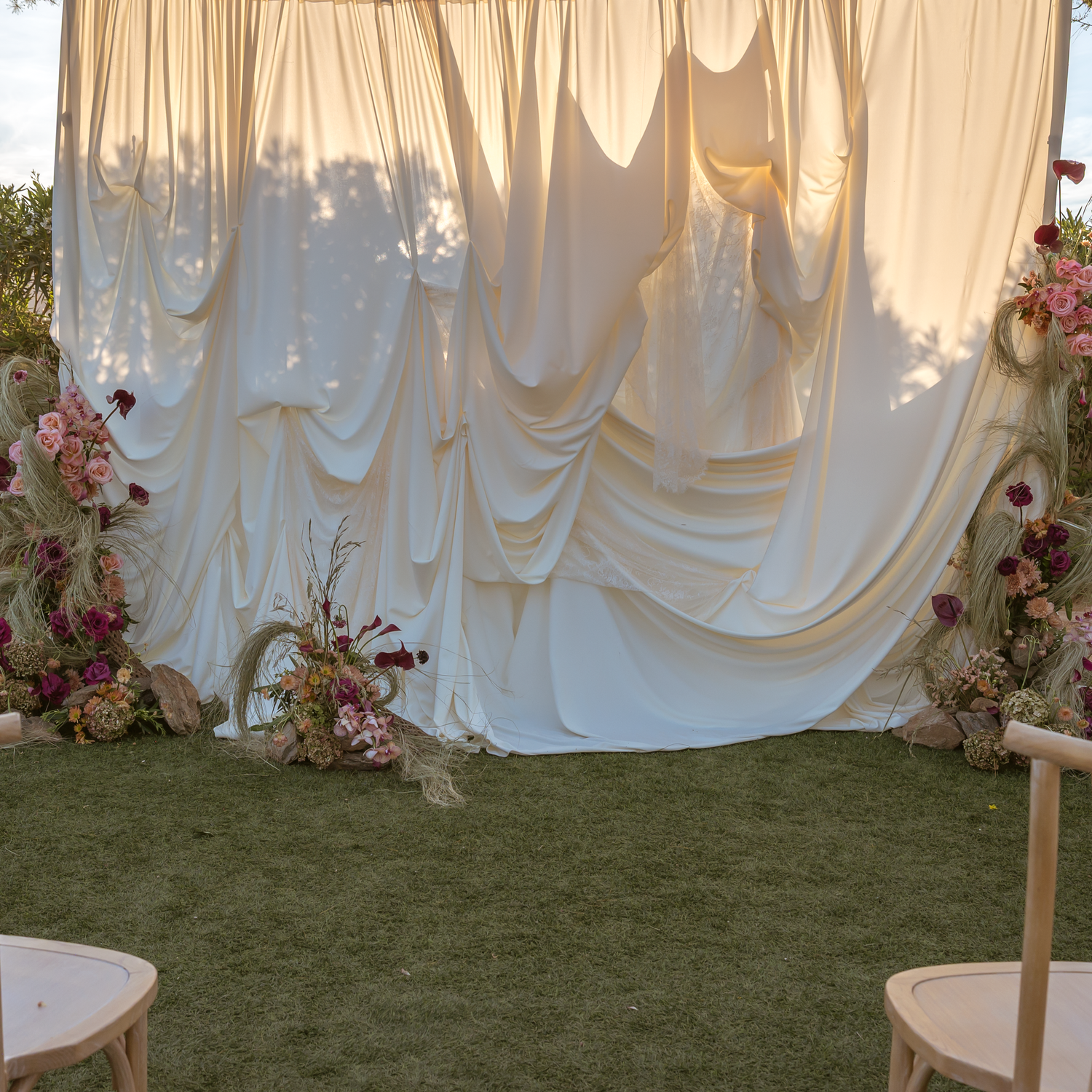 Elegant outdoor wedding ceremony backdrop with draped white fabric, pink floral arrangements, and lush green grass.