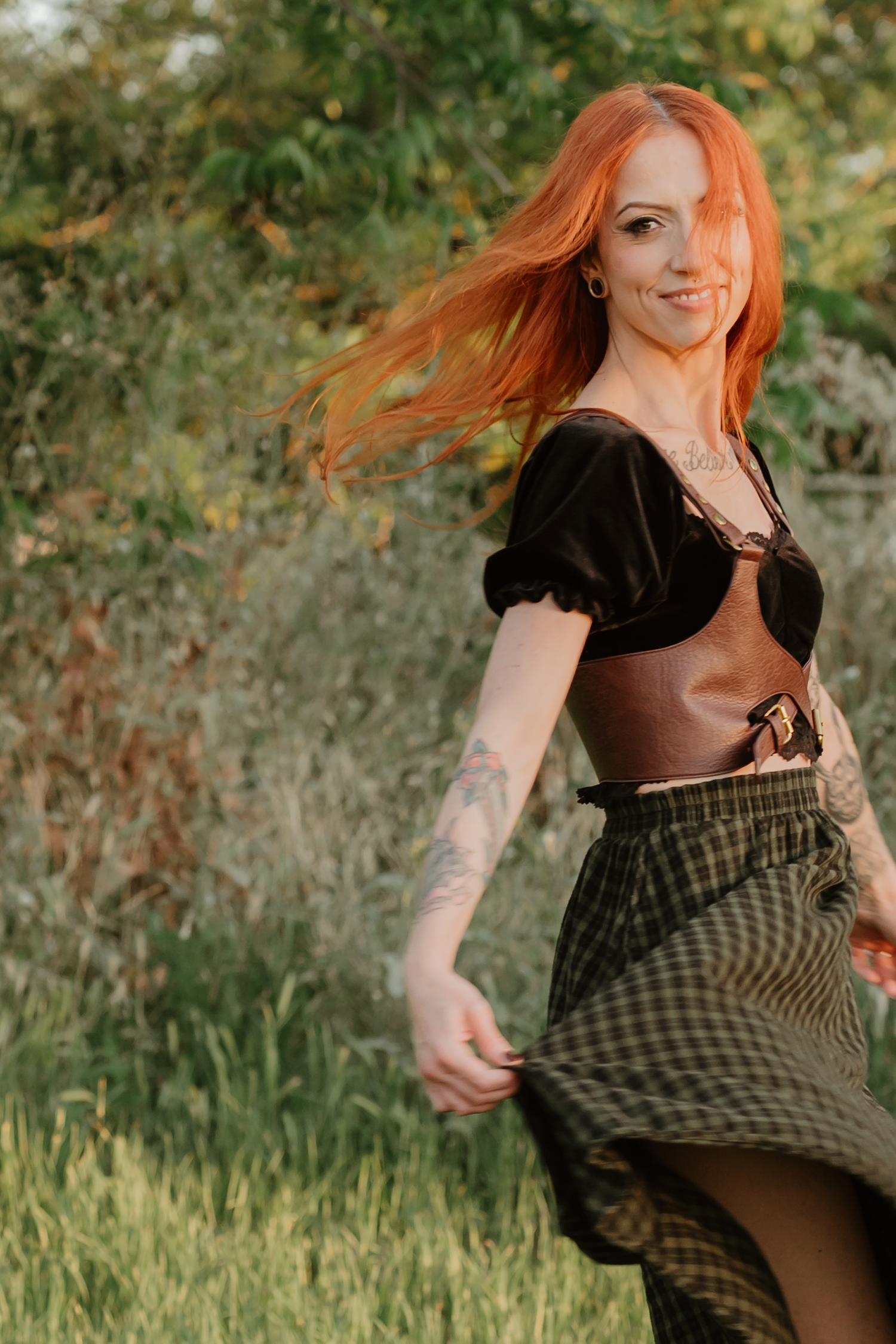 Red-haired woman with tattoos wearing a black top and plaid skirt, posing outdoors in a green natural setting.