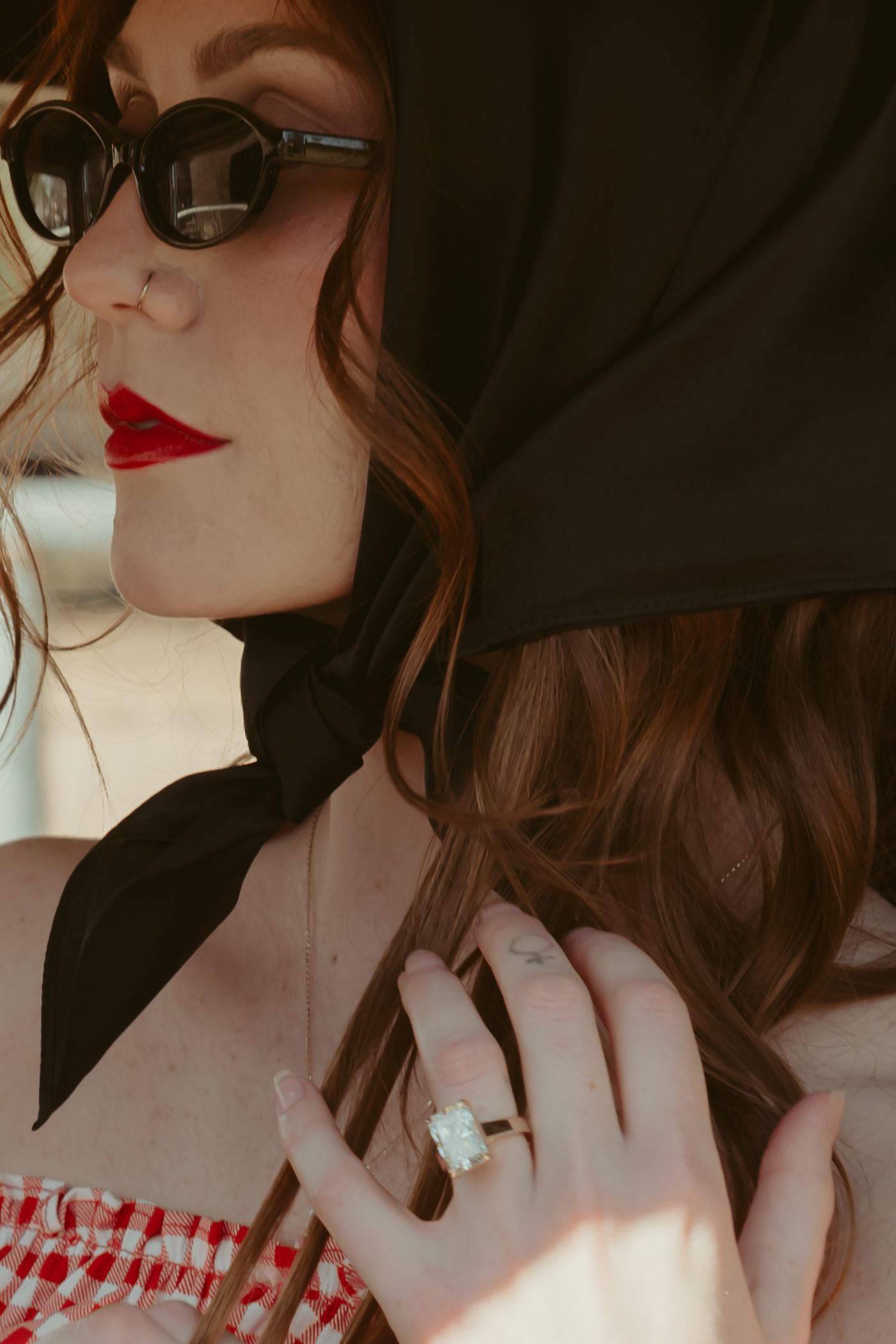 Stylish woman with red lips, black headscarf, sunglasses, and diamond ring wearing a red gingham top.