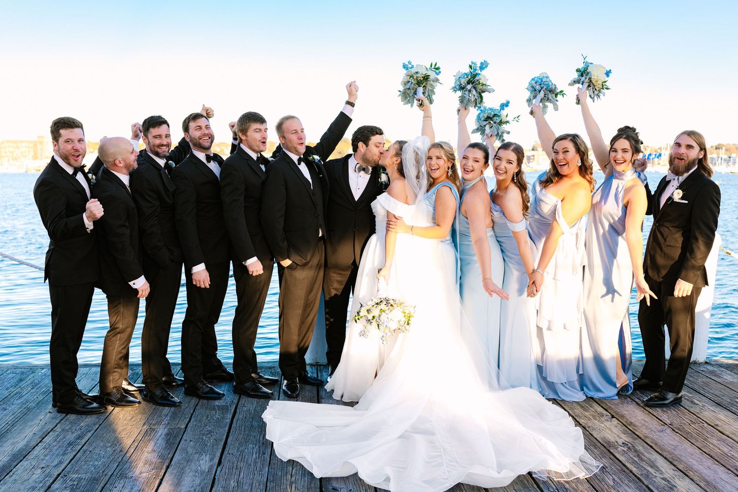 Wedding party in black suits and blue bridesmaid dresses pose together on wooden dock by waterfront at sunset at The Regatta Place in RI