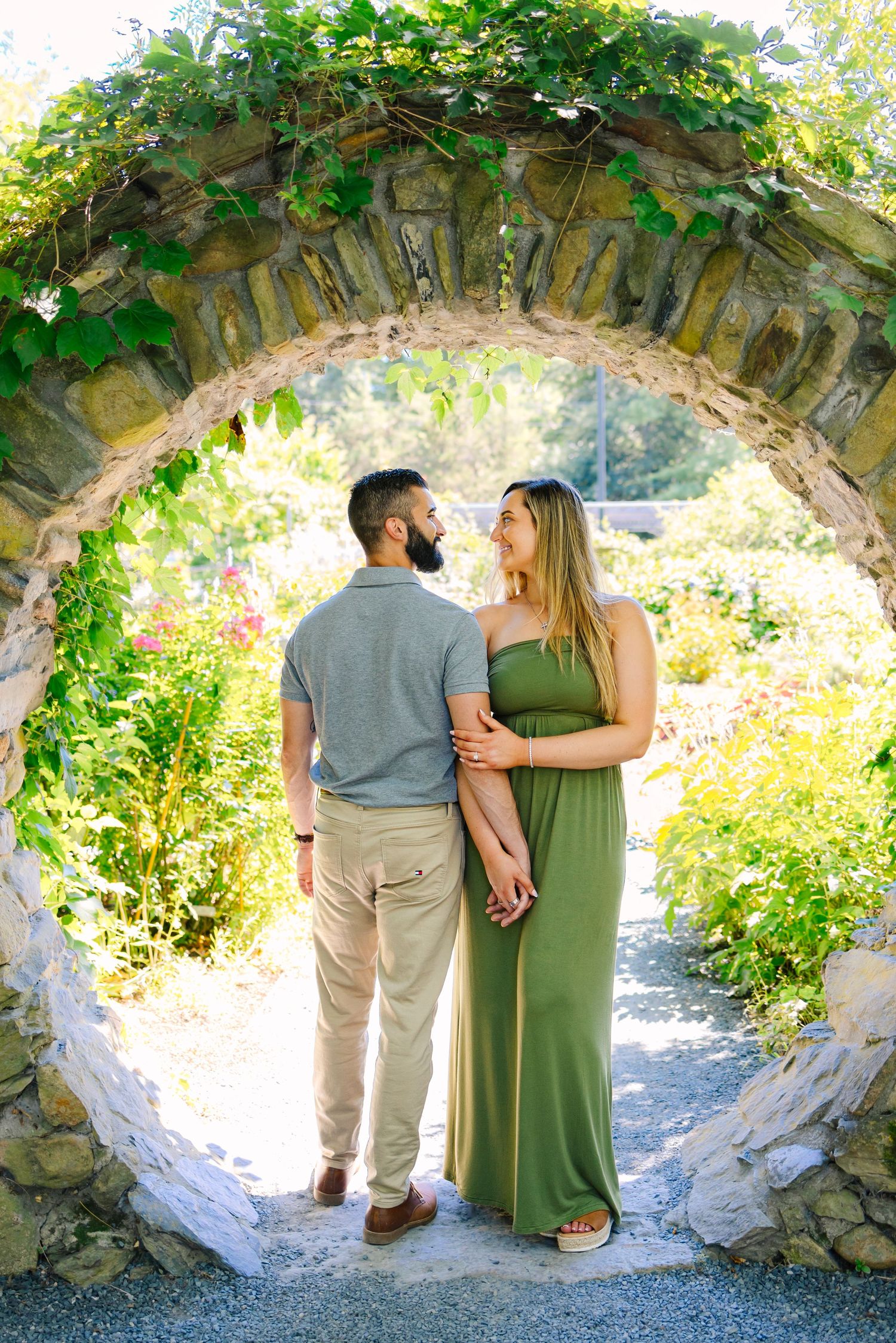 A romantic couple stands beneath a stone archway covered in green vines in a scenic garden setting at Blithewold Mansion in Bristol RI