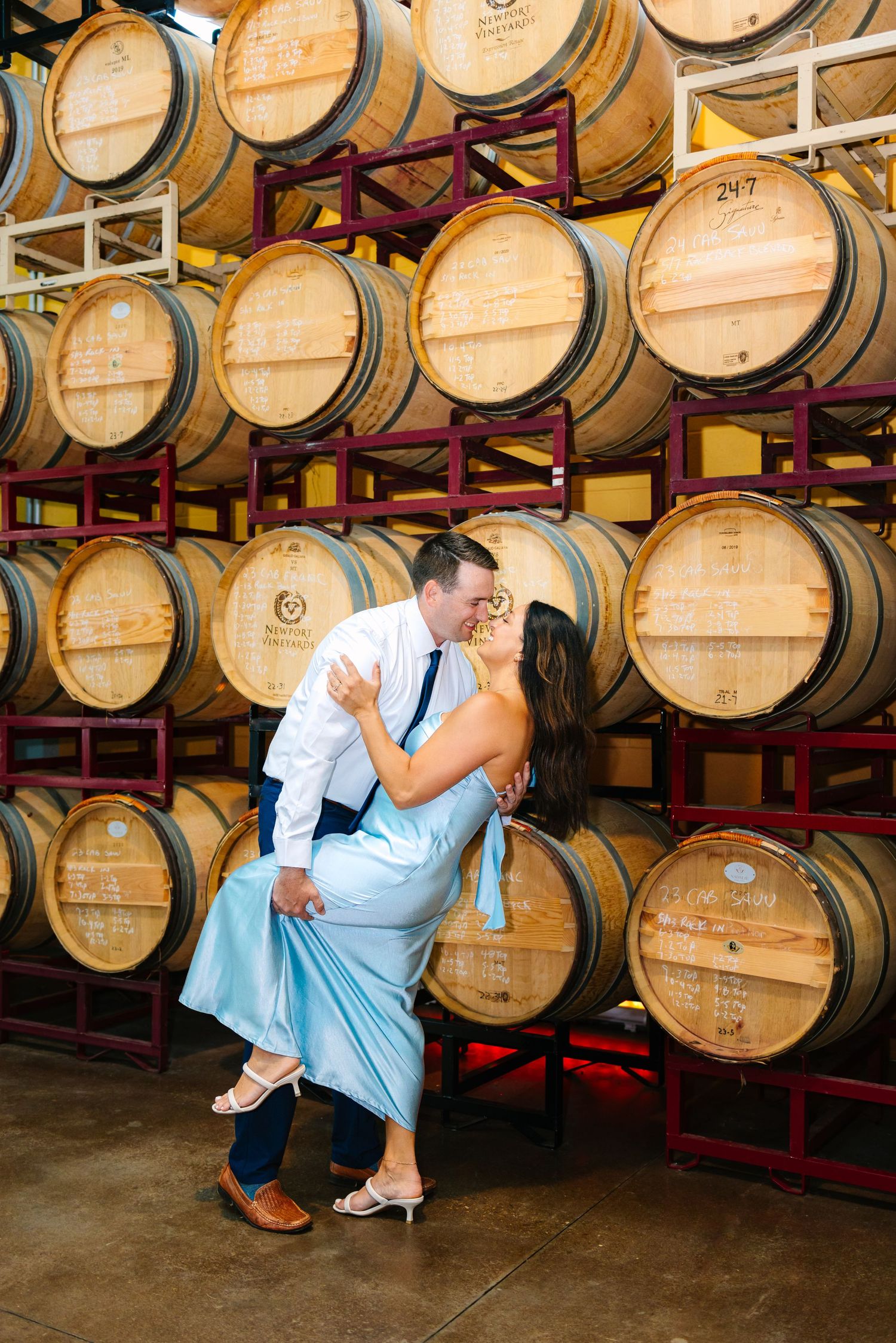 Wooden wine barrels stacked on metal racks in a rustic winery cellar with concrete floors with couple taking engagement photos in Newport RI