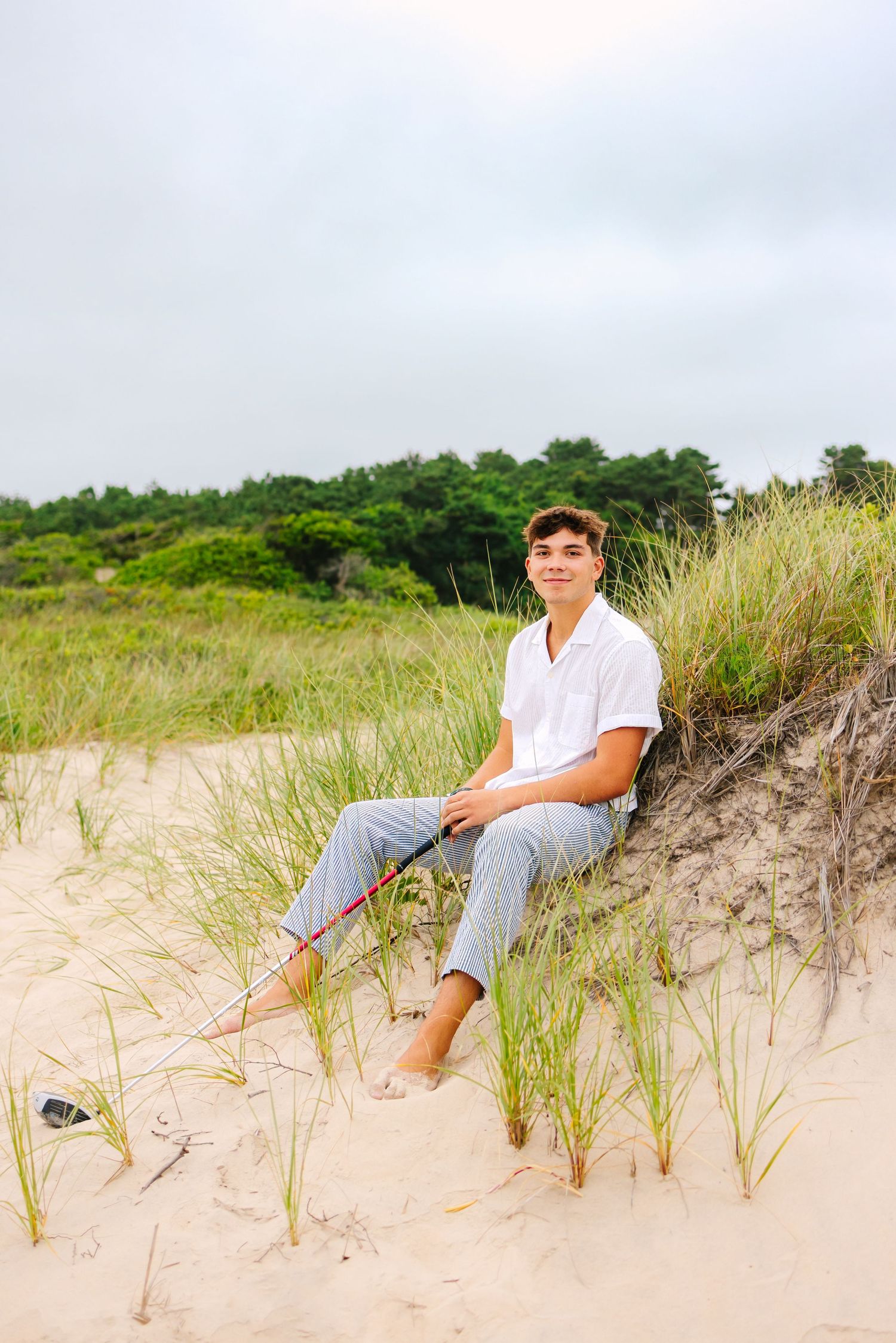 A high school senior posing with his golf club on the sand dunes at sunset golden hour for senior portraits at Horseneck Beach in MA