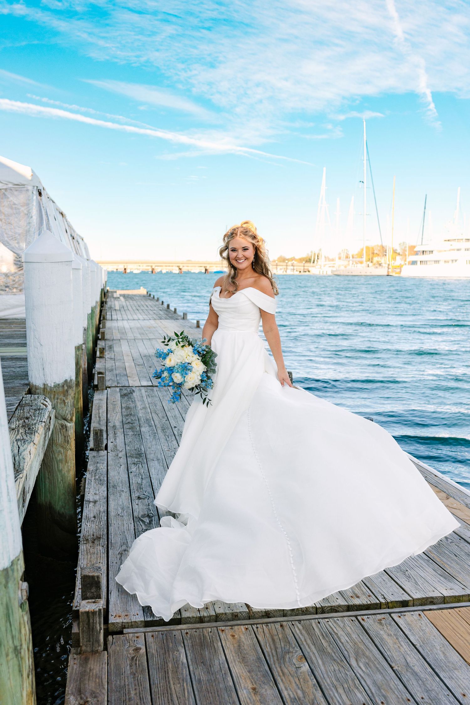 A bride in a flowing white gown twirls joyfully on a wooden dock by the ocean at sunset.