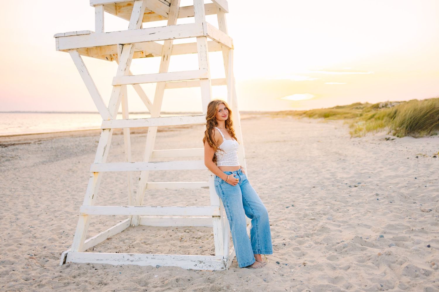 A high school senior leans against a lifeguard chair on the sand during sunset golden hour for senior photos at Horseneck Beach in MA