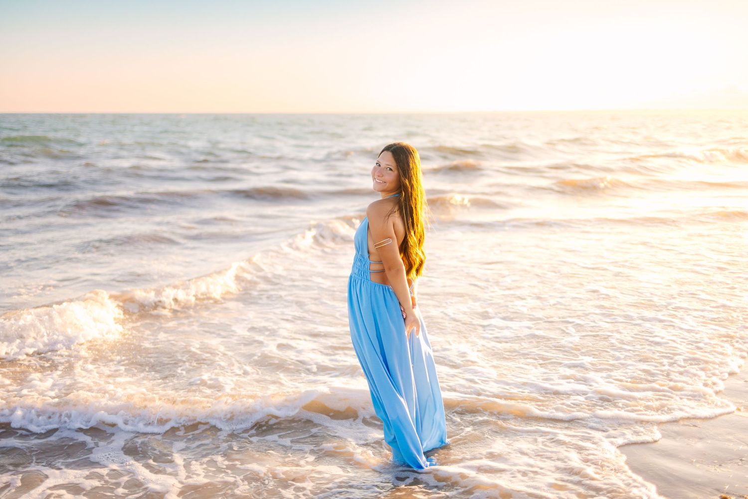 A high school senior in a flowing blue dress walks along the shoreline at sunset at Horseneck Beach in Westport Massachusetts