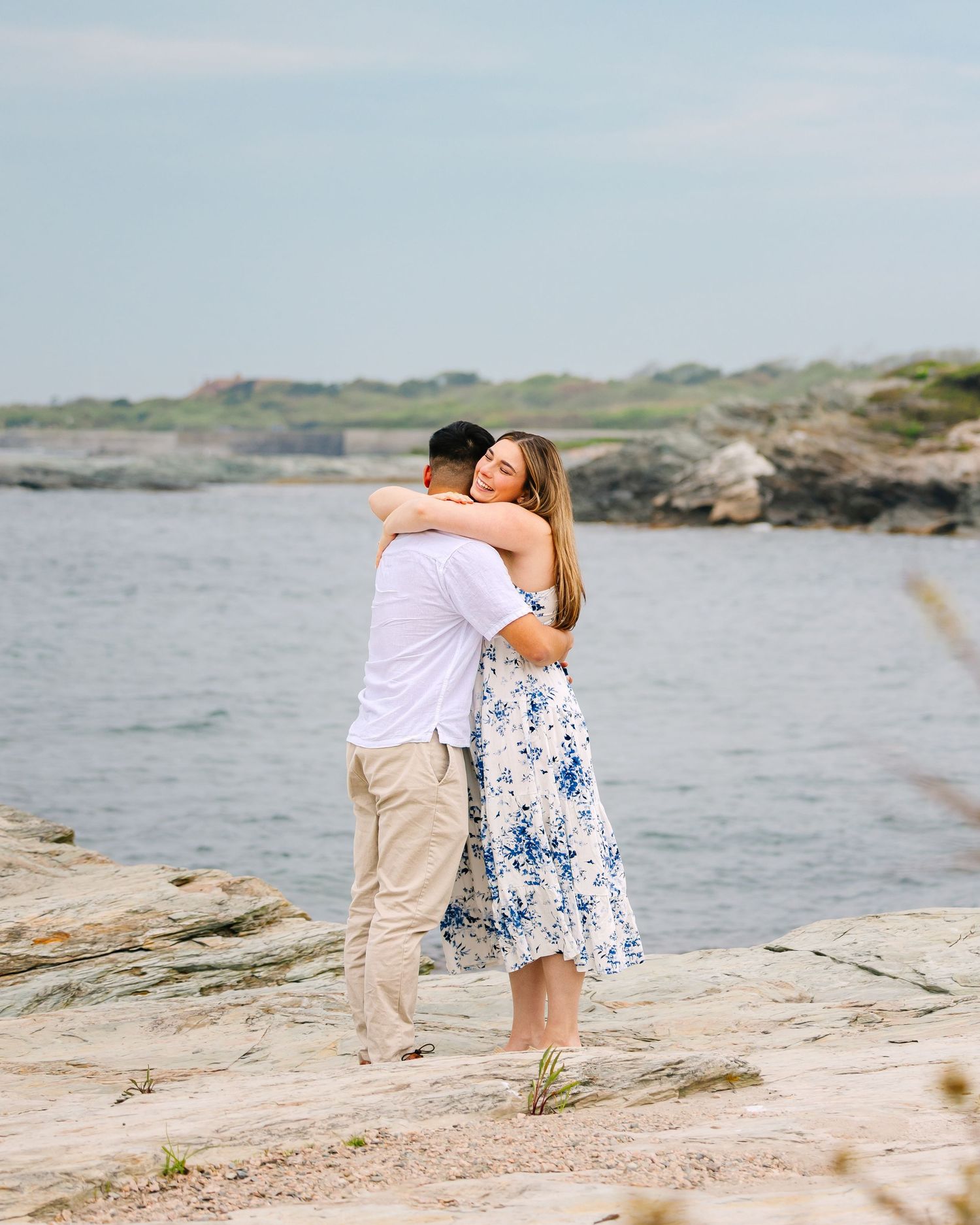 A romantic couple embraces on rocky coastal cliffs overlooking the ocean during a tranquil sunset on Ocean Drive in Newport Rhode Island