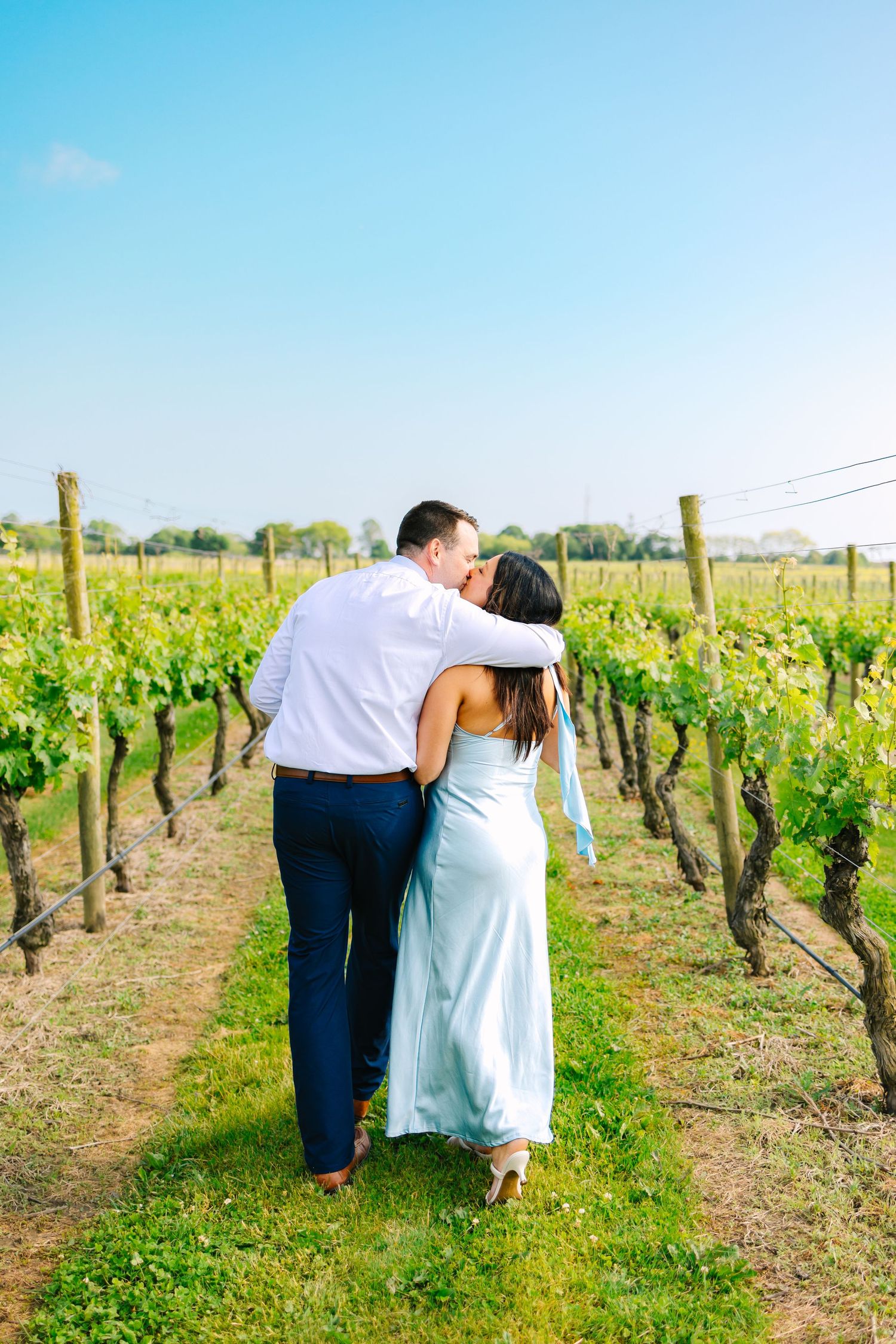 A couple walking arm in arm through lush green vineyard rows on a bright sunny day for engagement photos at Newport Vineyards in RI
