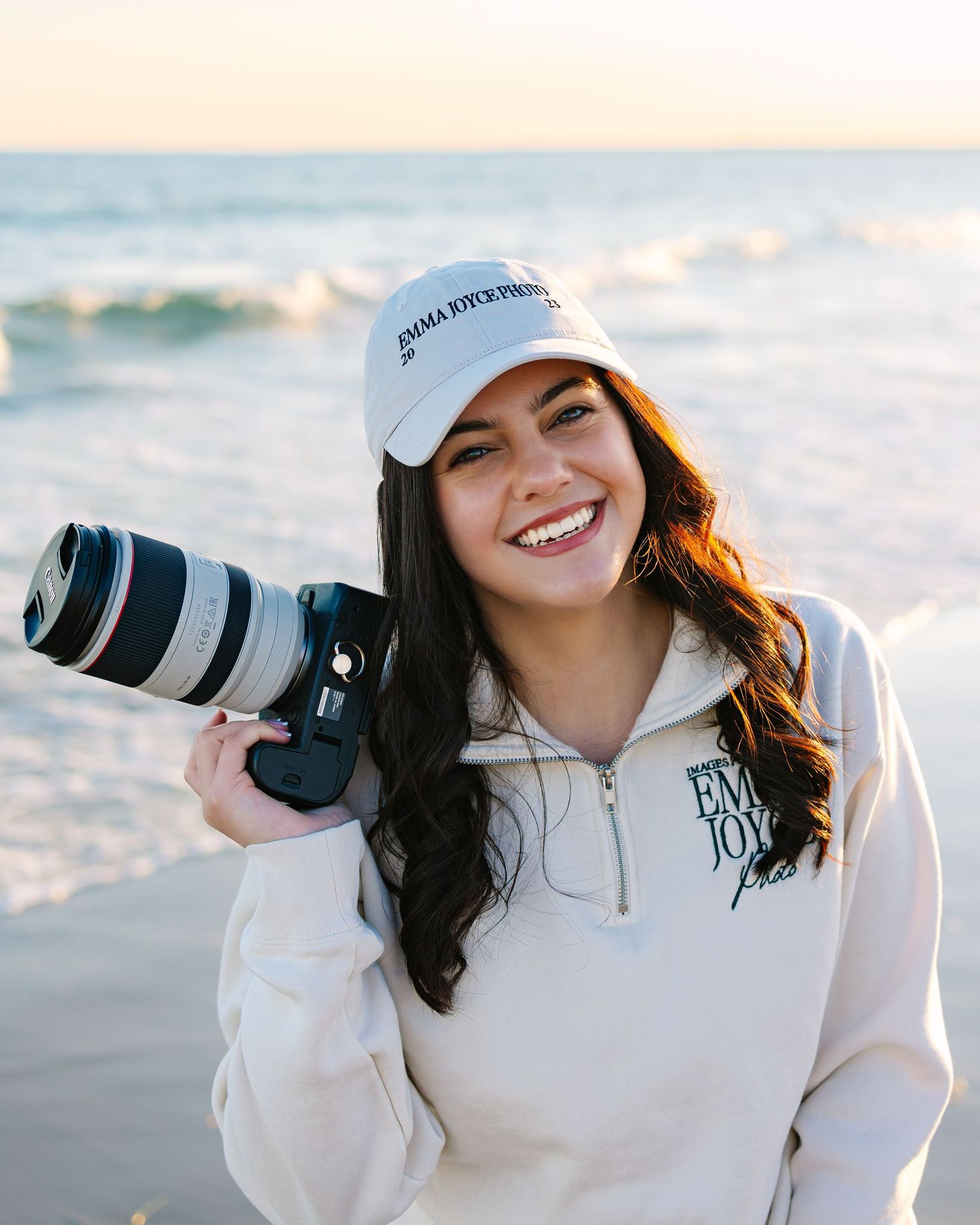 A photographer posing with her camera for branding photos by the shore at sunset golden hour at Horseneck Beach in Westport, Massachusetts