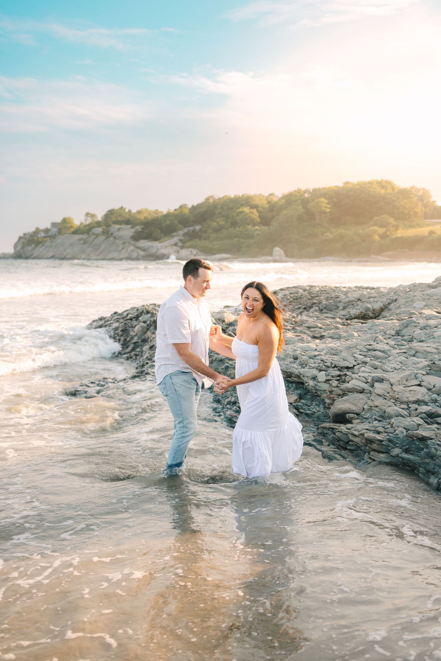A couple shares a romantic moment on a rocky beach at sunset as waves crash around them.