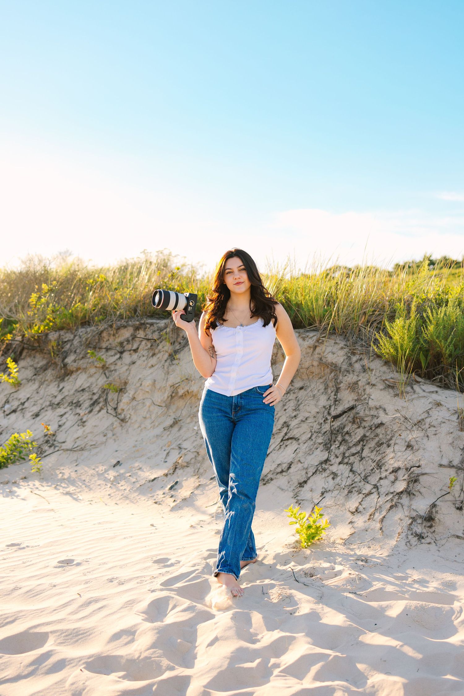 A photographer holds a professional camera while posing on sandy dunes with wildflowers and blue sky in the background.