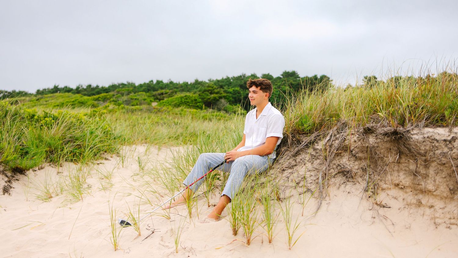 A high school senior sitting in sand dunes holding a golf club at sunset golden hour at Horseneck Beach in Westport Massachusetts
