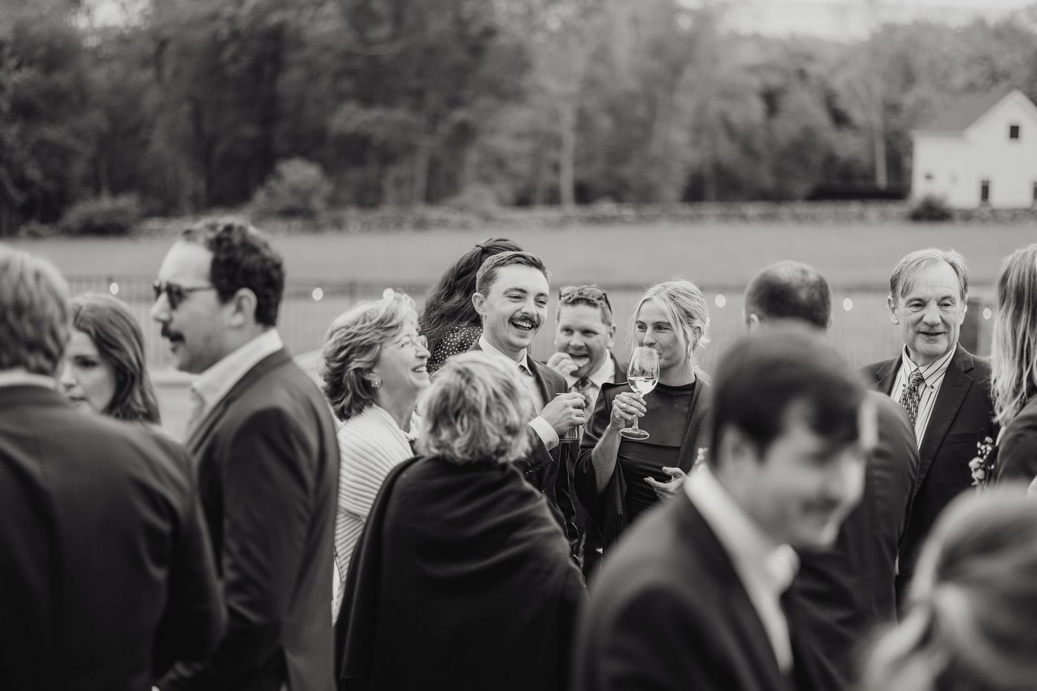 A black and white candid photograph of guests laughing and mingling at an outdoor wedding reception.