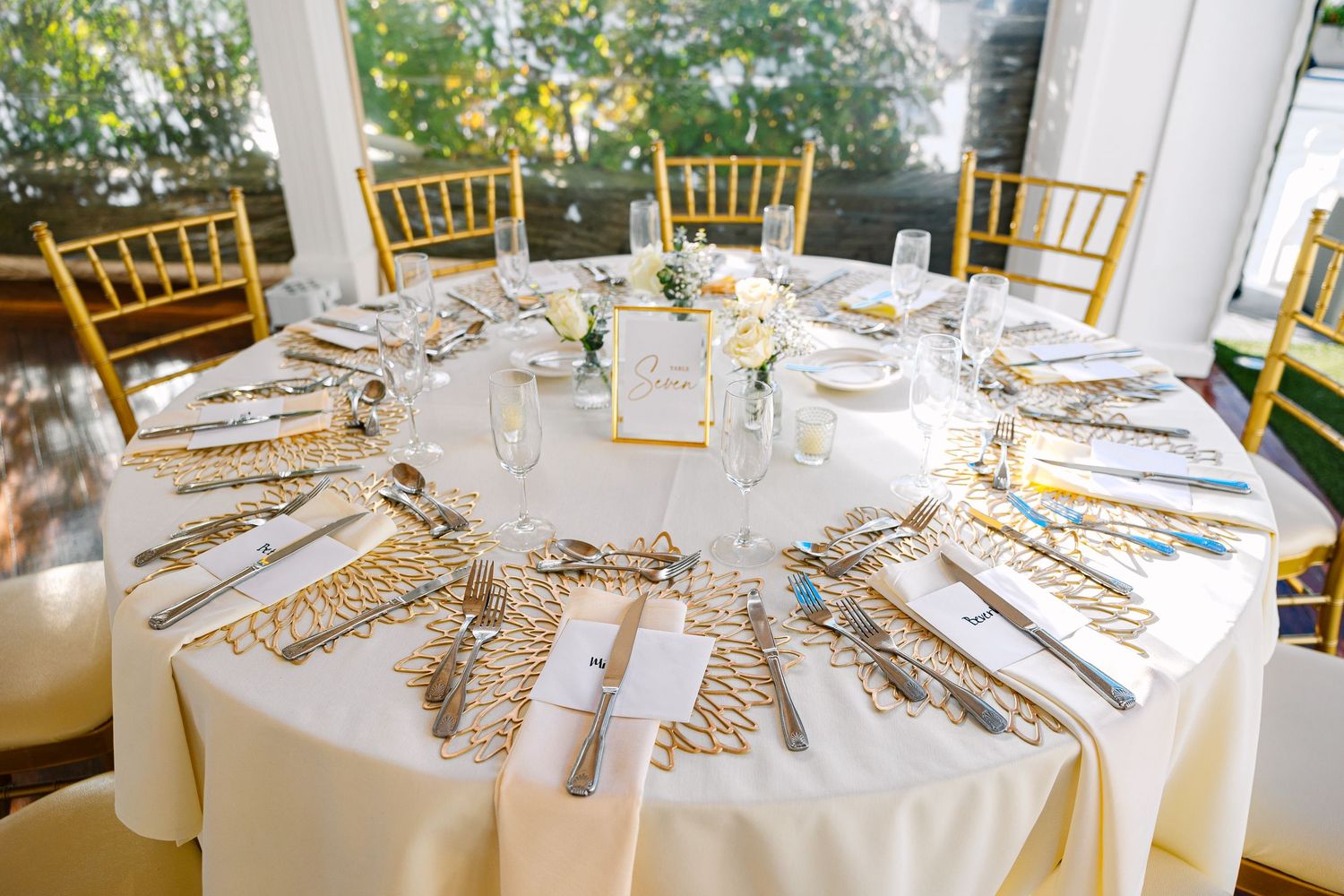 Elegant wedding reception table setting with gold chiavari chairs and white linens in a bright outdoor venue.