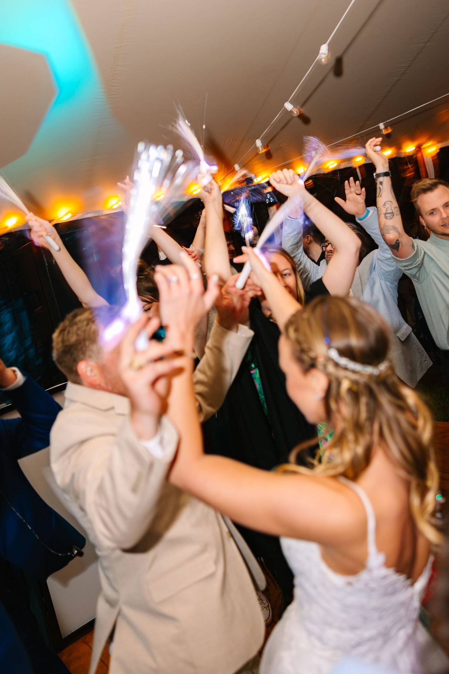 Energetic party crowd dancing with hands in the air under colorful blue and orange lighting at a nightclub event.