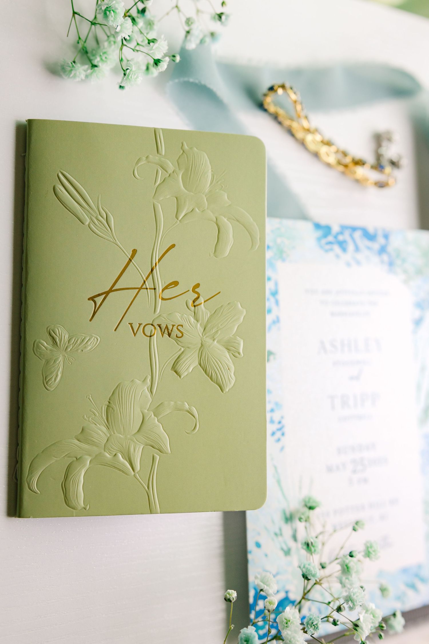 Elegant sage green vows book with embossed floral design and gold lettering displayed with delicate white flowers and blue details.