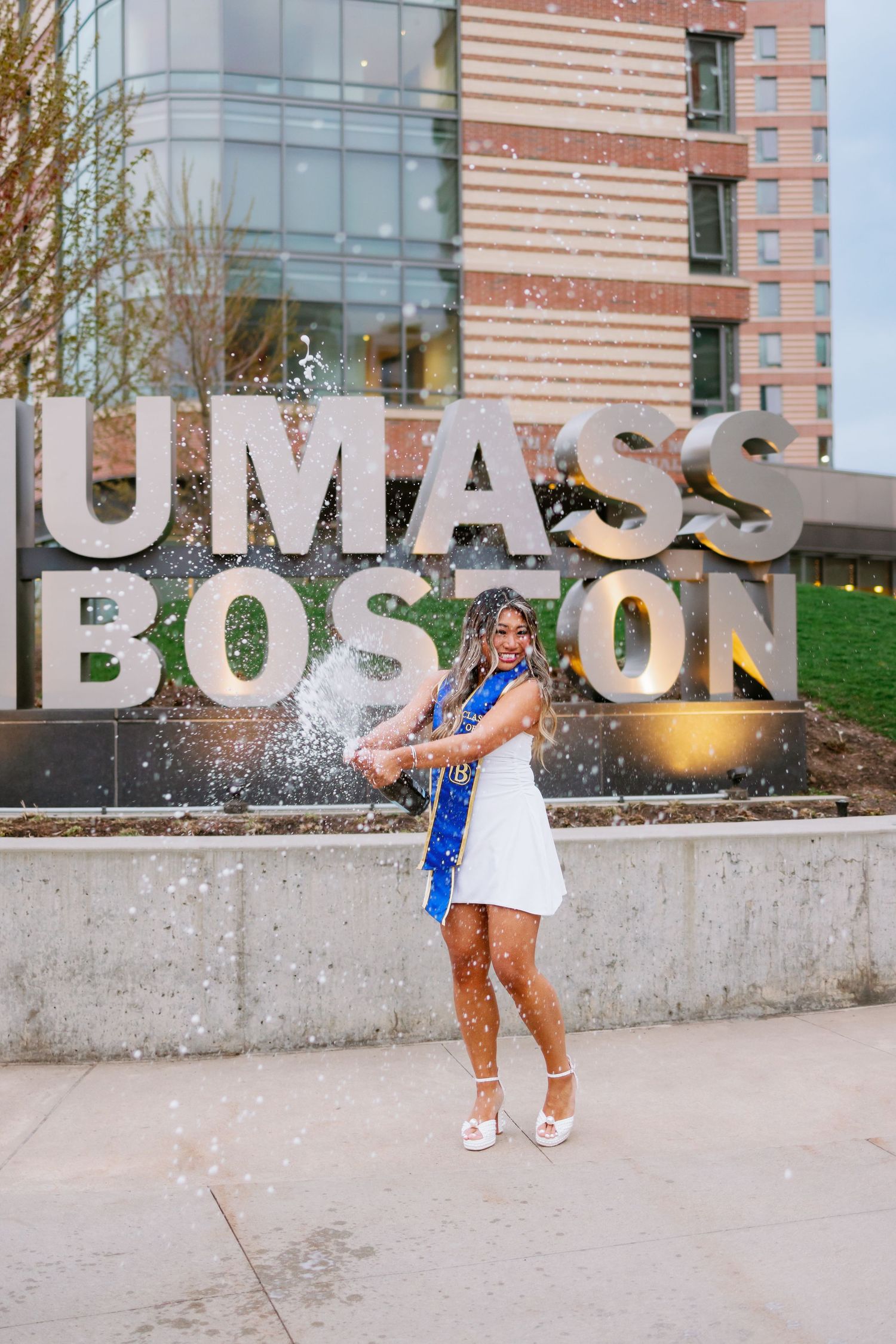 Graduate stands celebrating in front of large UMASS BOSTON letters on campus wearing white dress and blue stole.