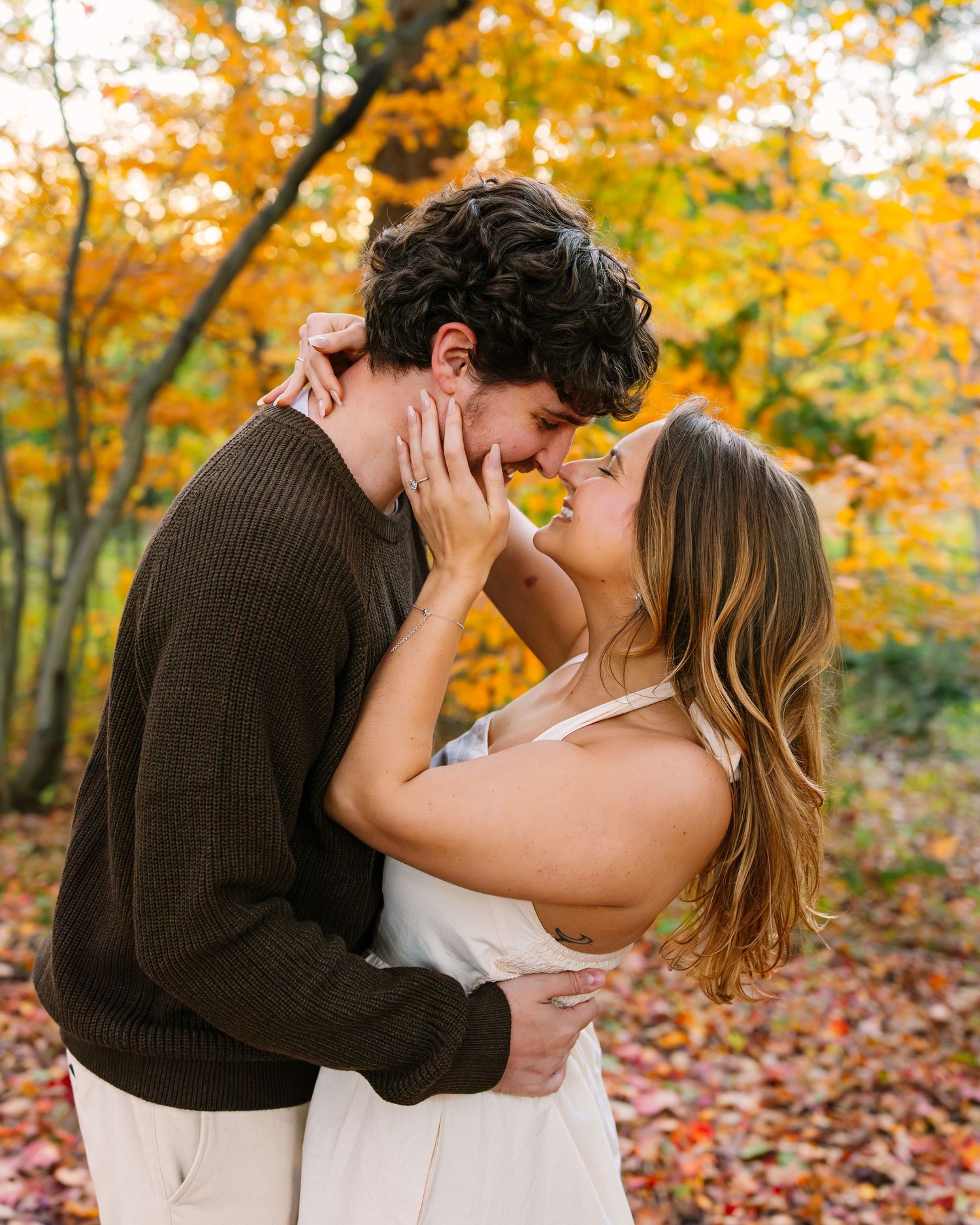 A romantic couple embraces and kisses in a vibrant autumn forest with golden leaves at Arnold Arboretum in Boston MA for engagement photos.